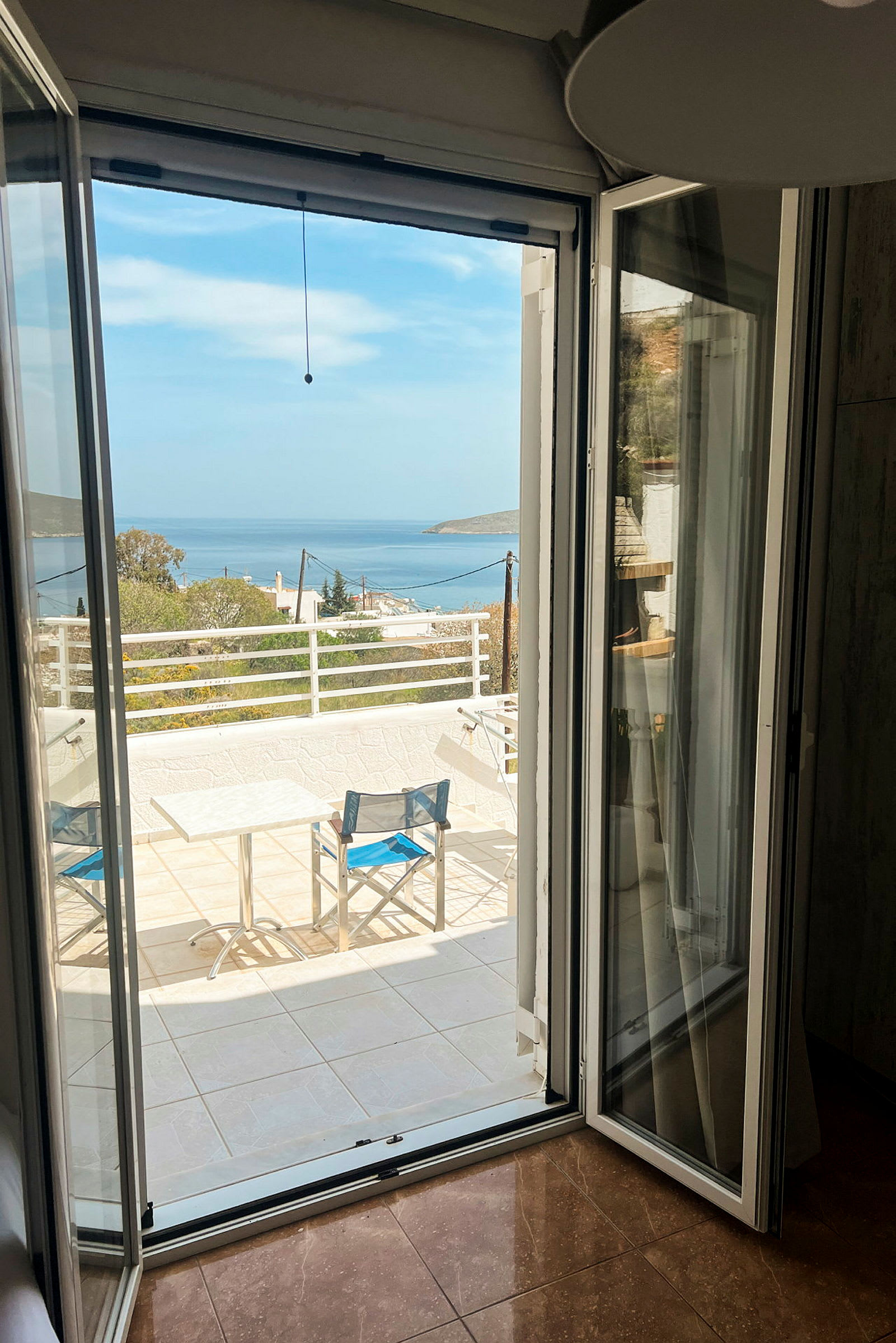 Udsigt fra et åbent vindue mod en terrasse med bord og stole samt havet i baggrunden. (AI-genereret tekst)