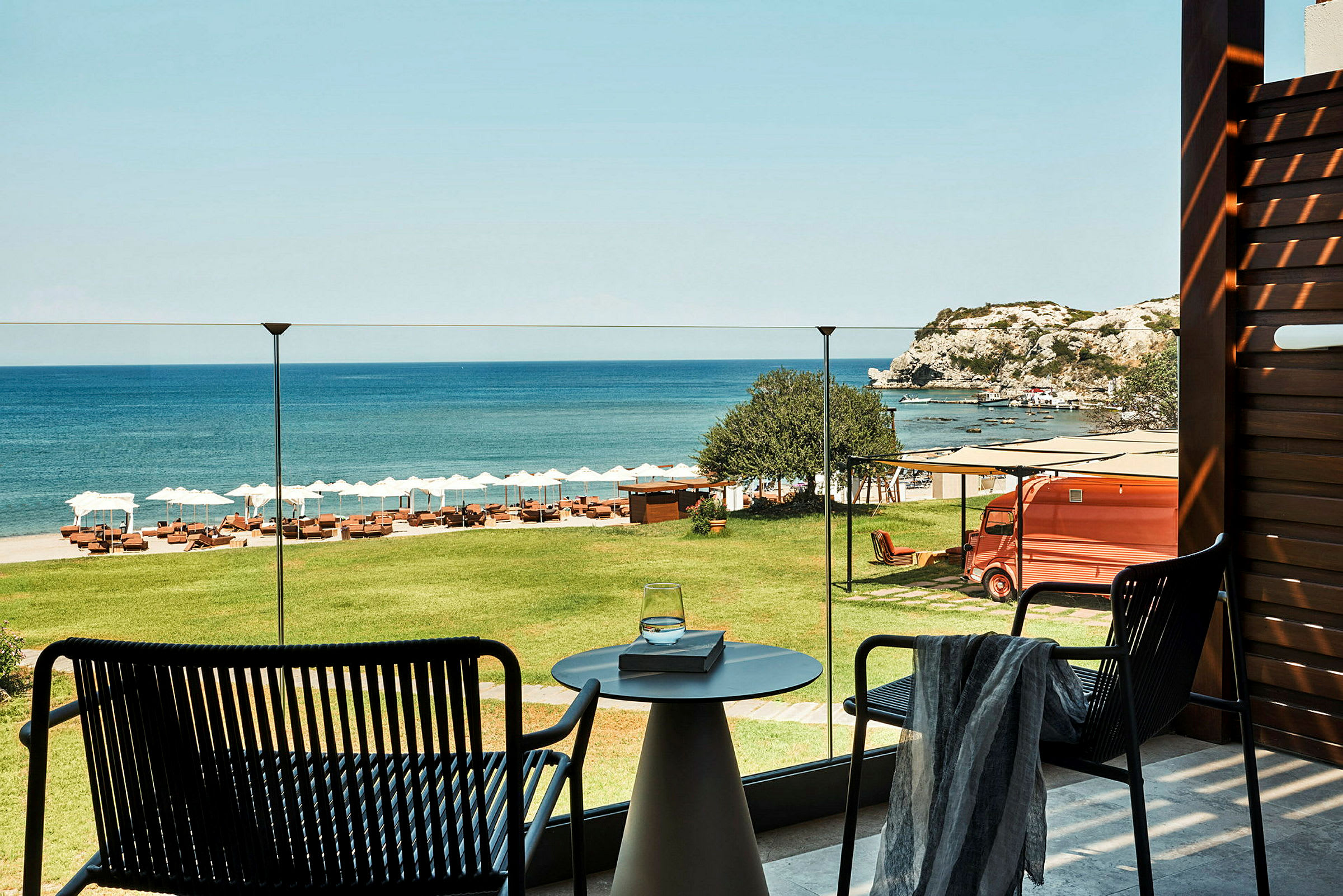 Udsigt fra en terrasse over grønt græs, en strand med liggestole og parasoller samt havet og klipper i baggrunden. (AI-genereret tekst)