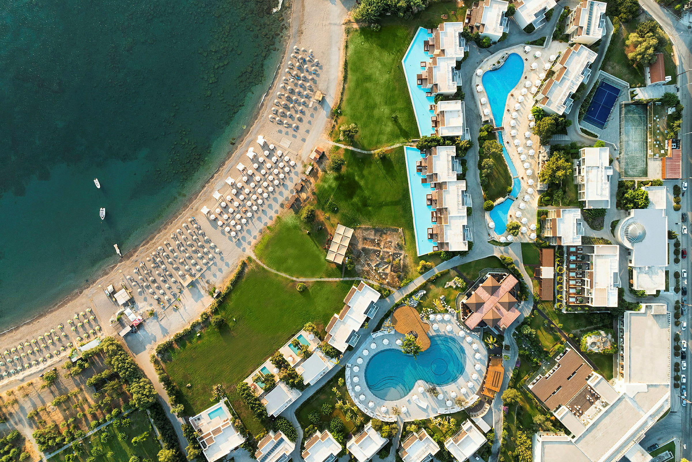 Luftfoto af en ferieby med bygninger, swimmingpools og en strand med liggestole. (AI-genereret tekst)