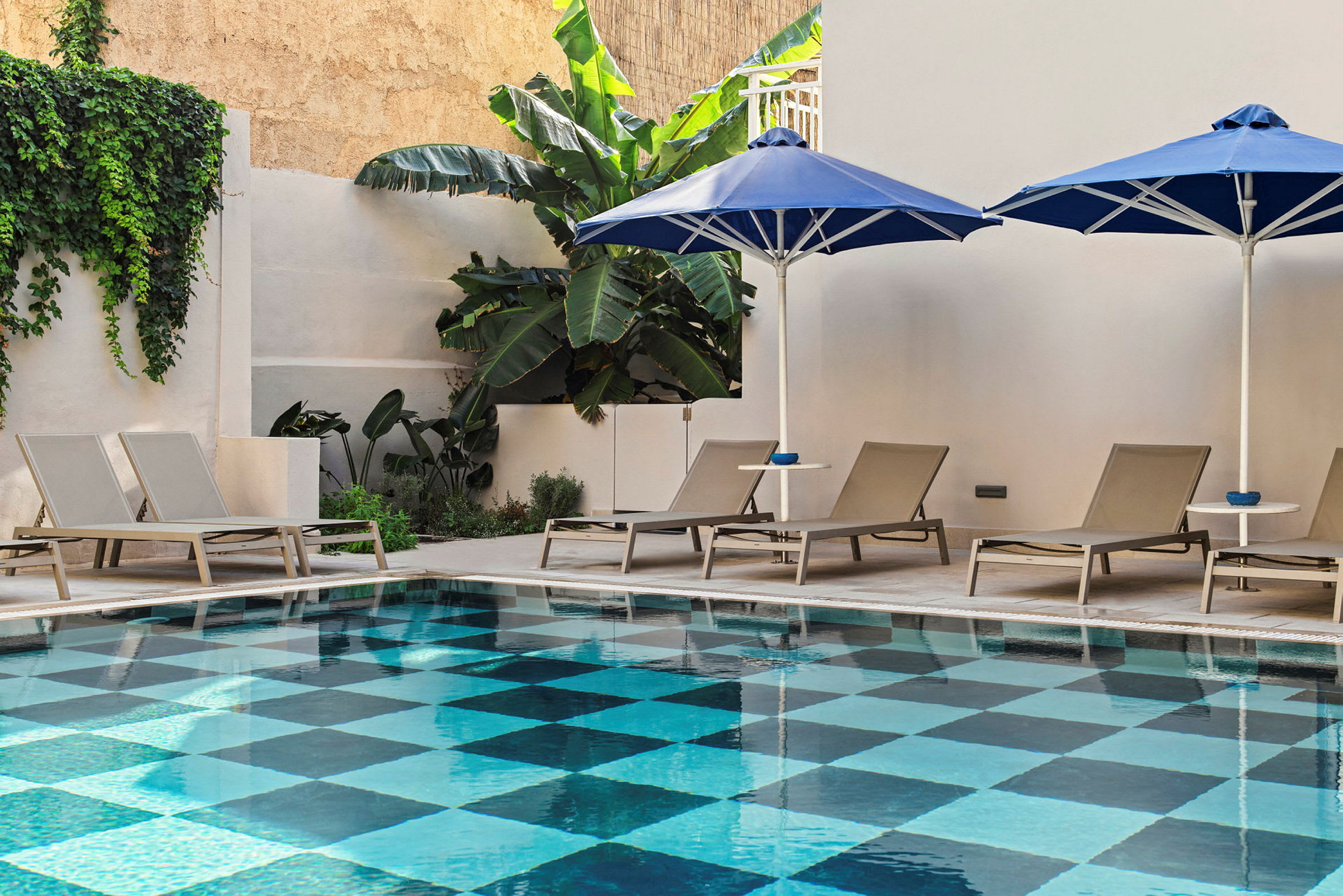 Rektangulær pool med skakternet bund omgivet af liggestole og parasoller. (AI-genereret tekst)