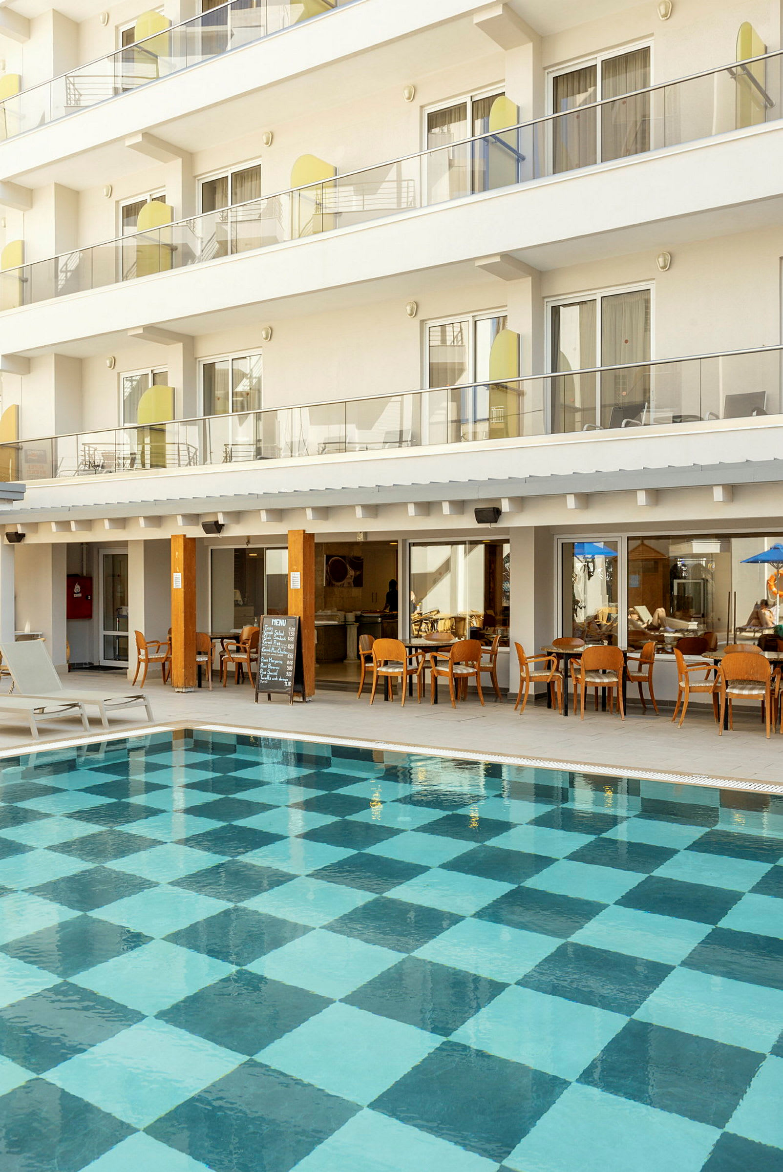 Udendørs pool med ternet bundmønster foran hotelbygning med altaner og restaurant. (AI-genereret tekst)