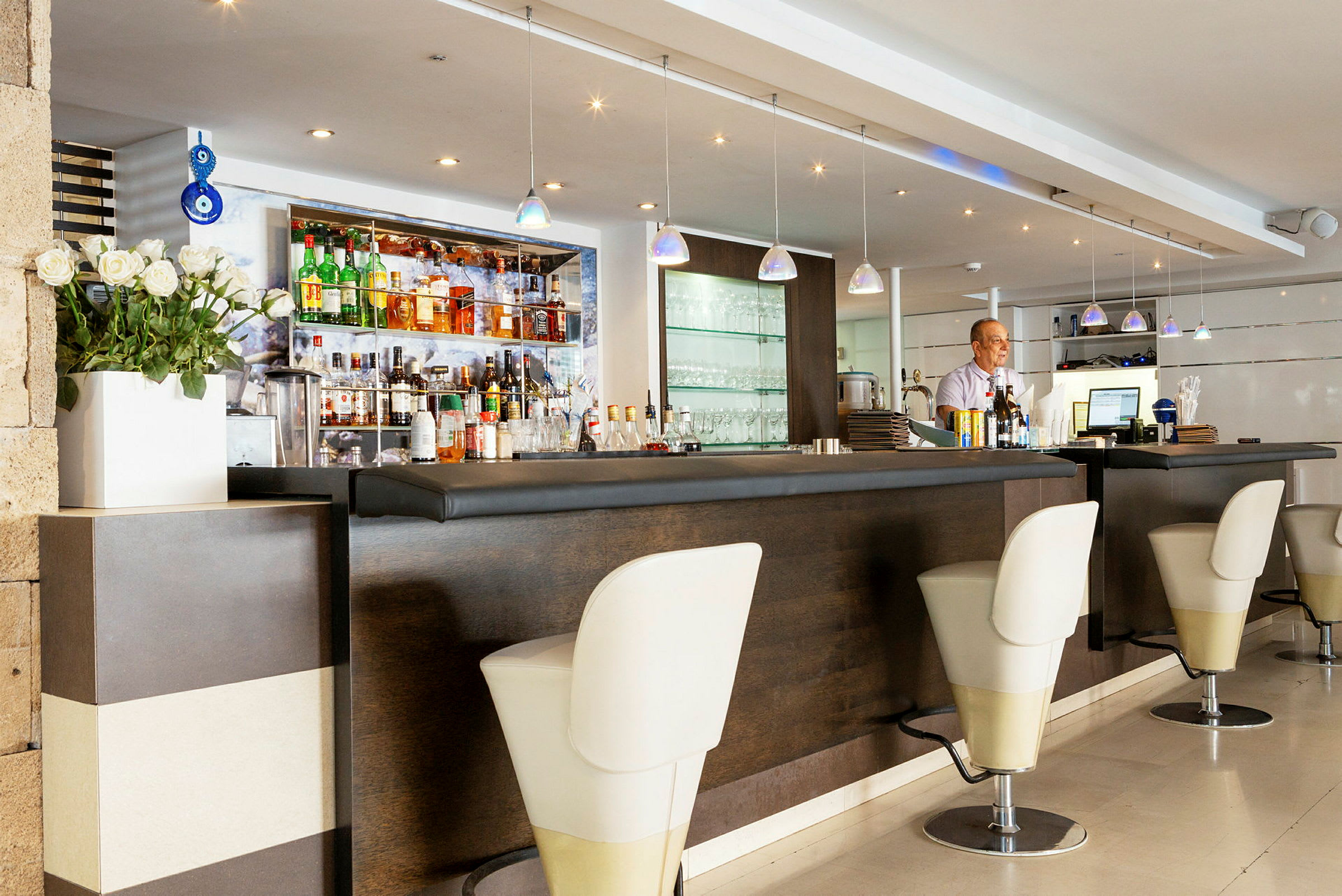 En hotelbar med elegant indretning inklusive et siddeområde med høje stole og flasker samt glas på en hylde. (AI-genereret tekst)