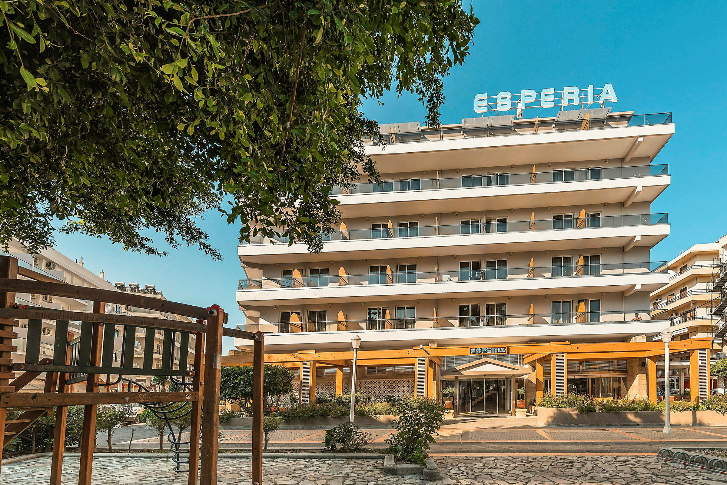 Hotelbygning med flere etager og et skilt med teksten 'Esperia' øverst. Der er legeredskaber i forgrunden. (AI-genereret tekst)