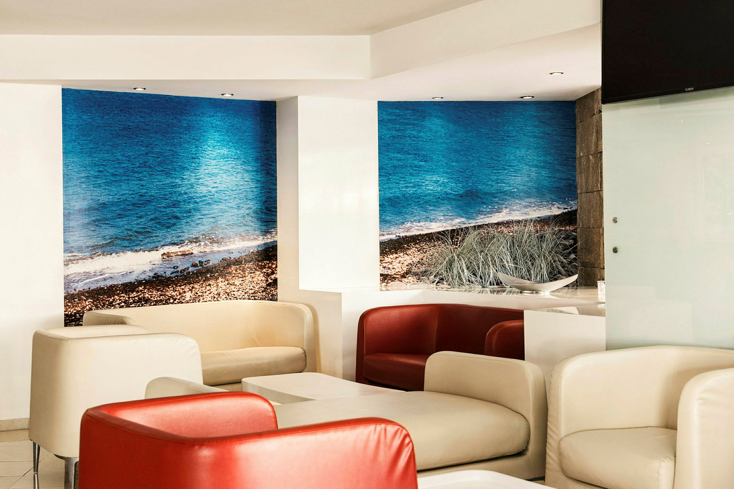 En moderne lounge med komfortable stole i hvidt og rødt samt borde. Væggene har en fototapet med motiv af en strand med blåt hav. (AI-genereret tekst)
