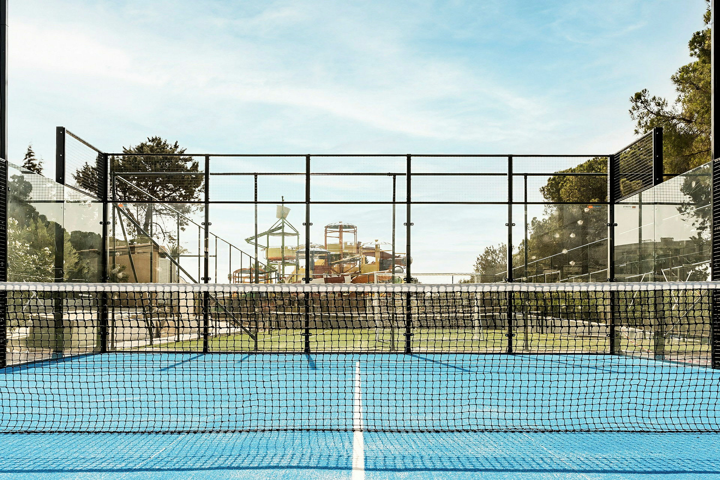 En padelbane med blå overflade og net i forgrunden, med en vandpark i baggrunden. (AI-genereret tekst)