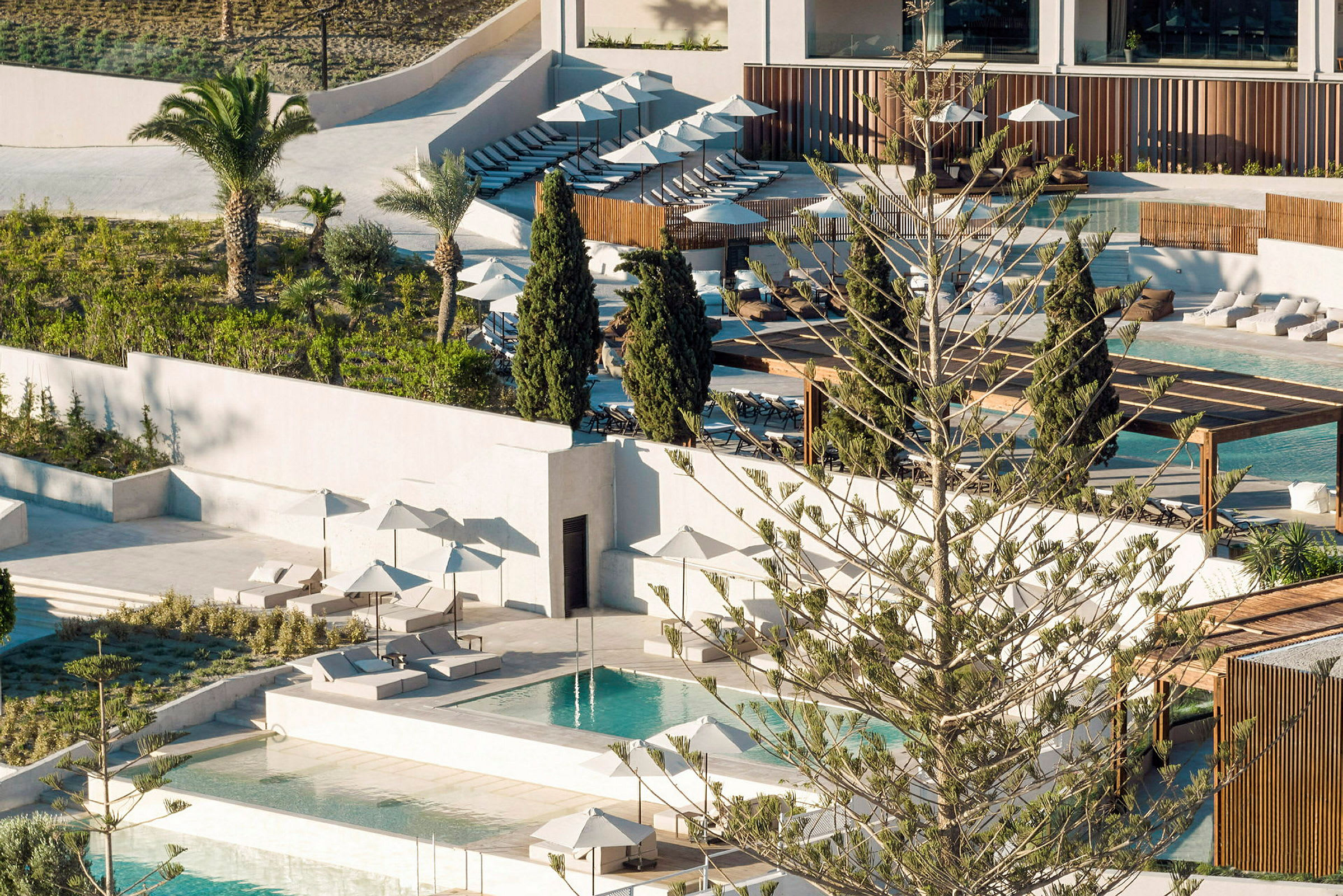 Udsigt over et hotelområde med pools, solsenge og parasoller, omgivet af grønne områder. (AI-genereret tekst)