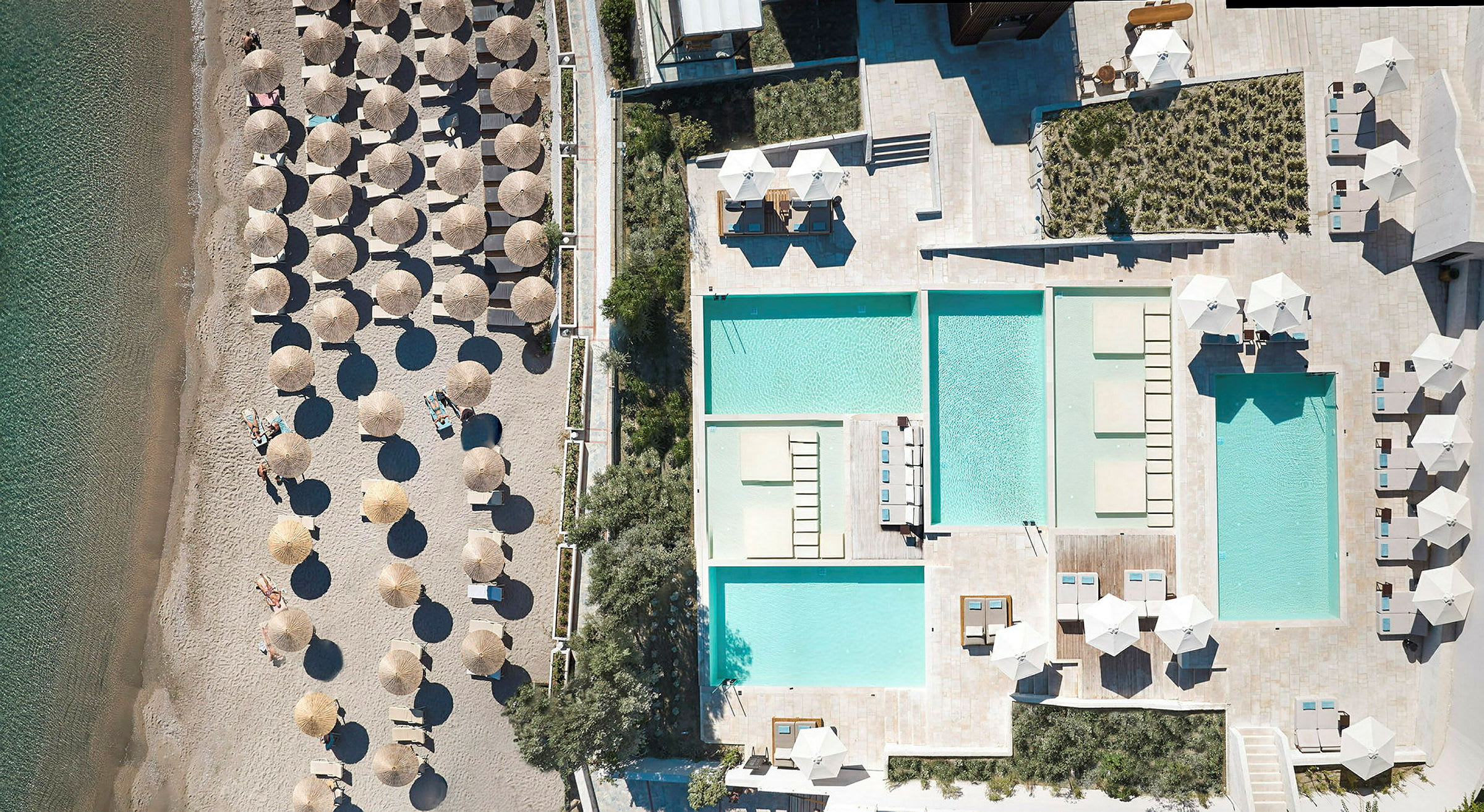 Luftfoto af en strand med parasoller og liggestole ved siden af en facilitet med flere swimmingpools. (AI-genereret tekst)