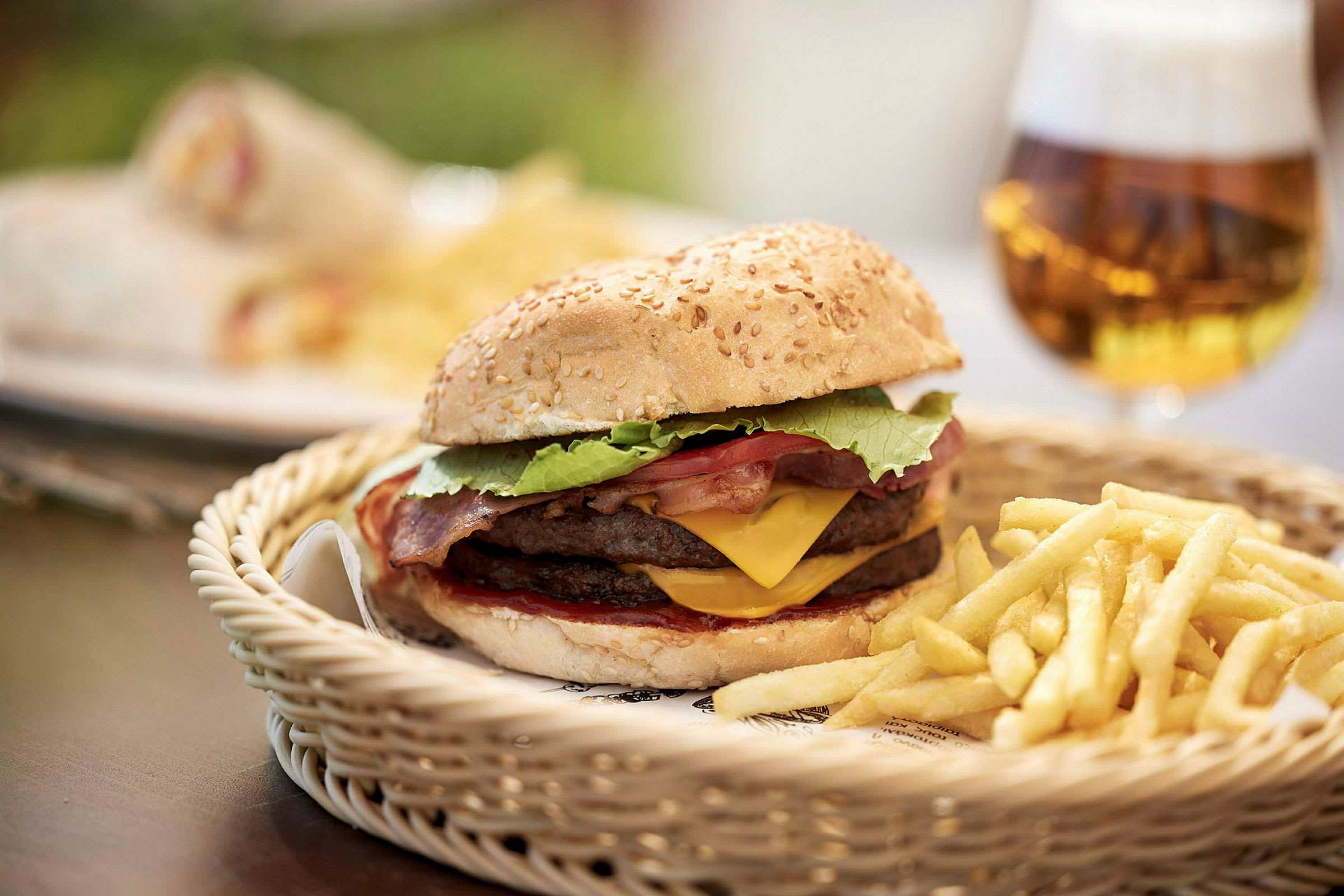Hamburger med ost og bacon, serveret med pommes frites i en kurv, samt en øl i baggrunden. (AI-genereret tekst)