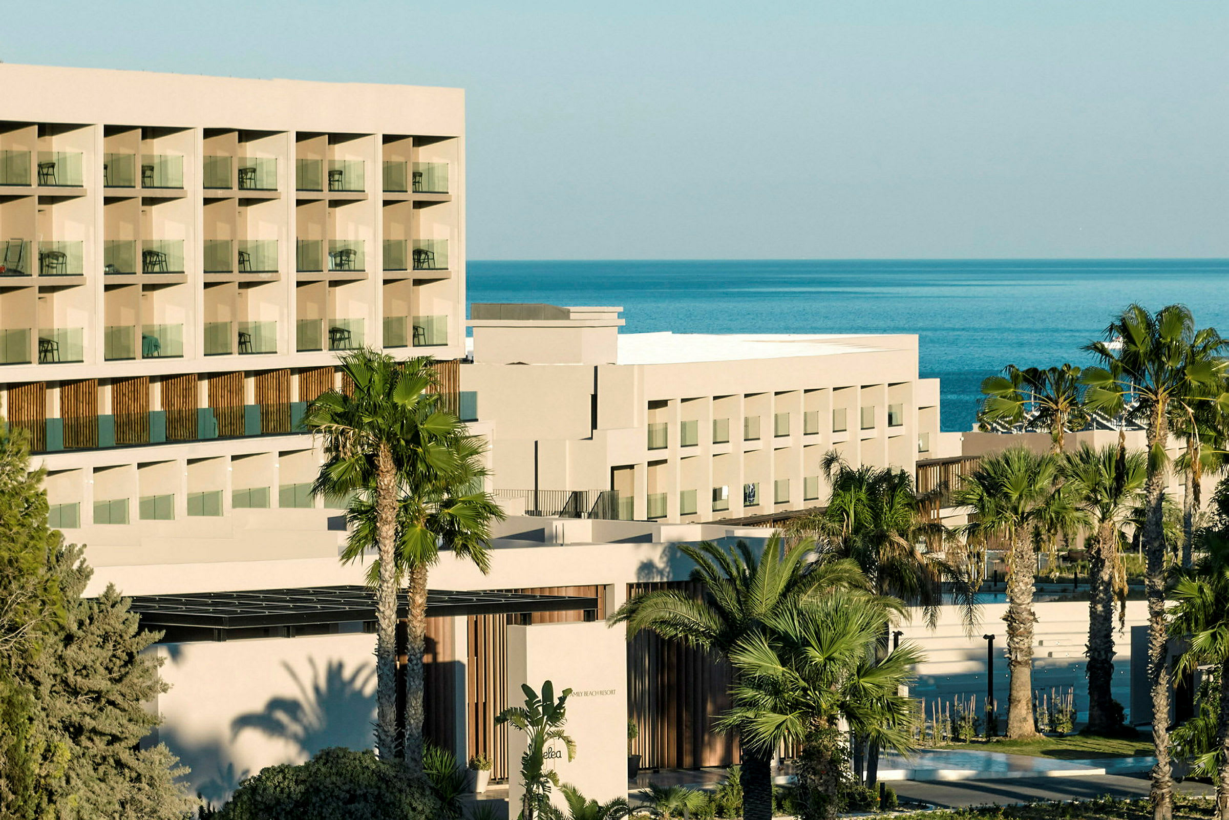 Billede af en moderne hotelbygning med flere etager og altaner, omgivet af palmer og med udsigt over havet. (AI-genereret tekst)