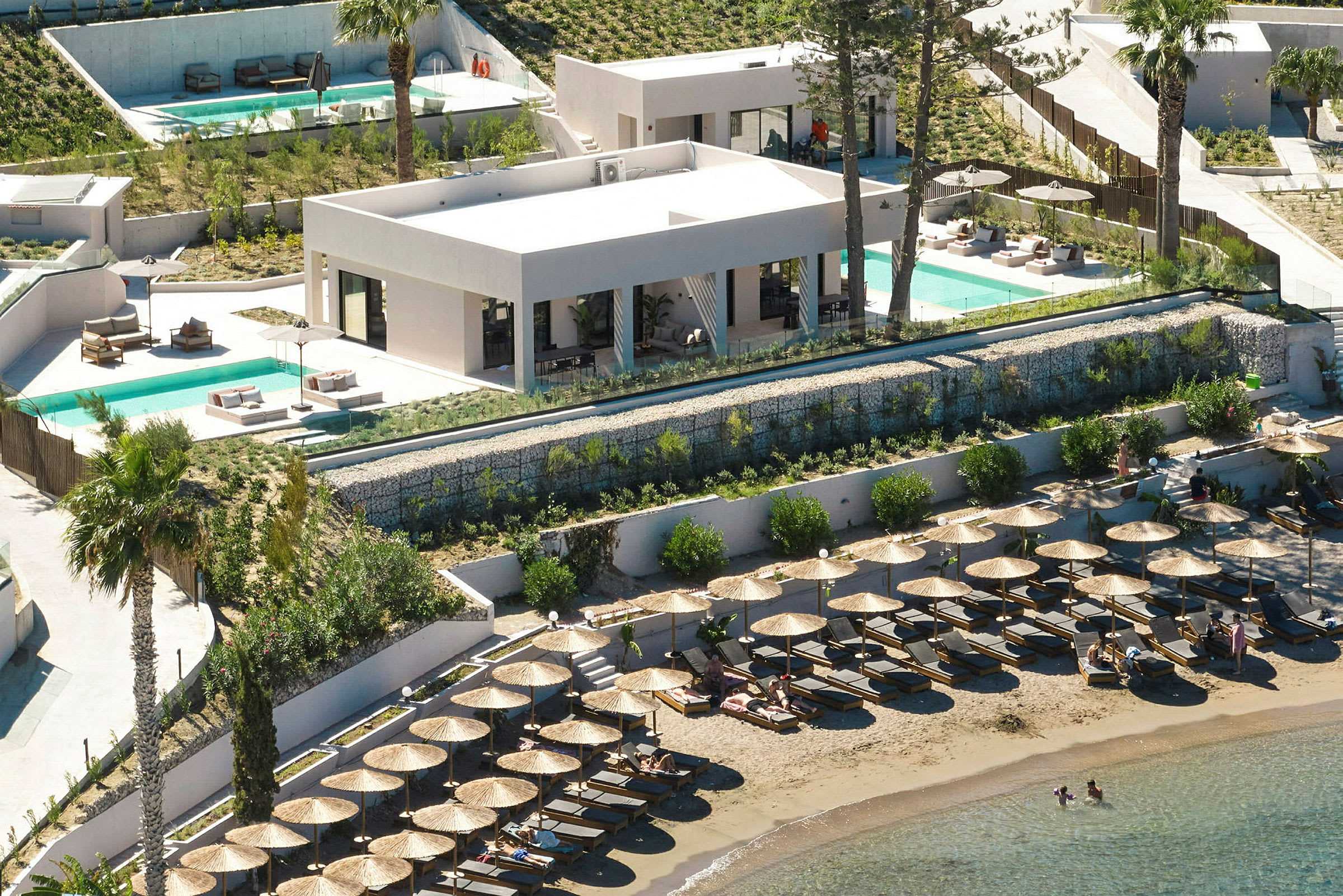Udsigt over en strand med liggestole og parasoller, bagved en bygning med pool og terrasse. (AI-genereret tekst)
