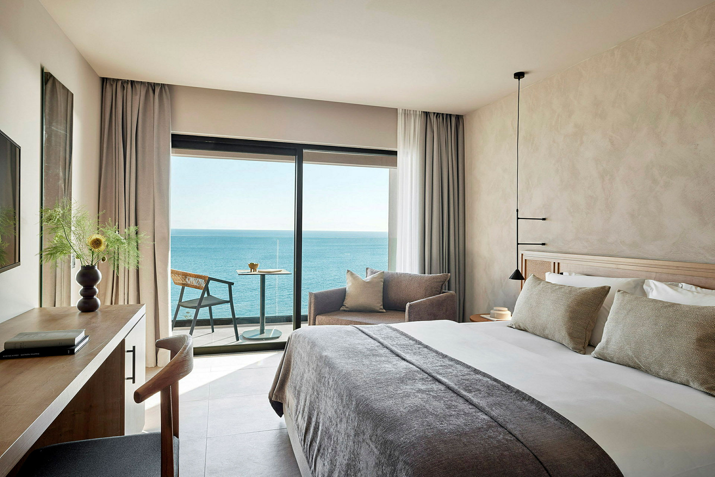 Et moderne hotelværelse med en stor seng, sofasektion og skrivebord samt en balkon med udsigt over havet. (AI-genereret tekst)