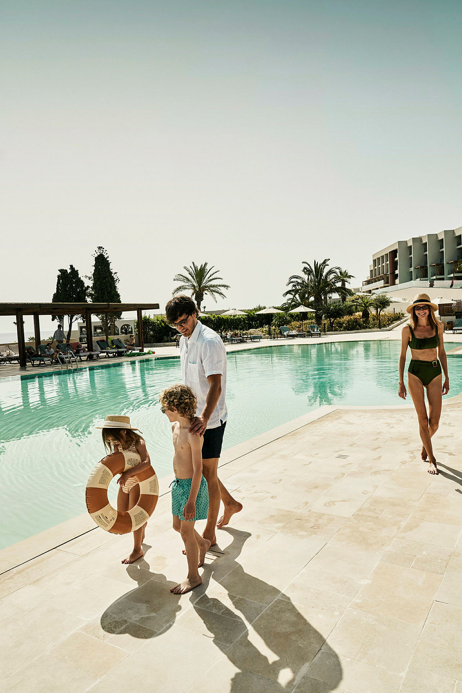 En familie nær en swimmingpool med en bygning og palmer i baggrunden. (AI-genereret tekst)