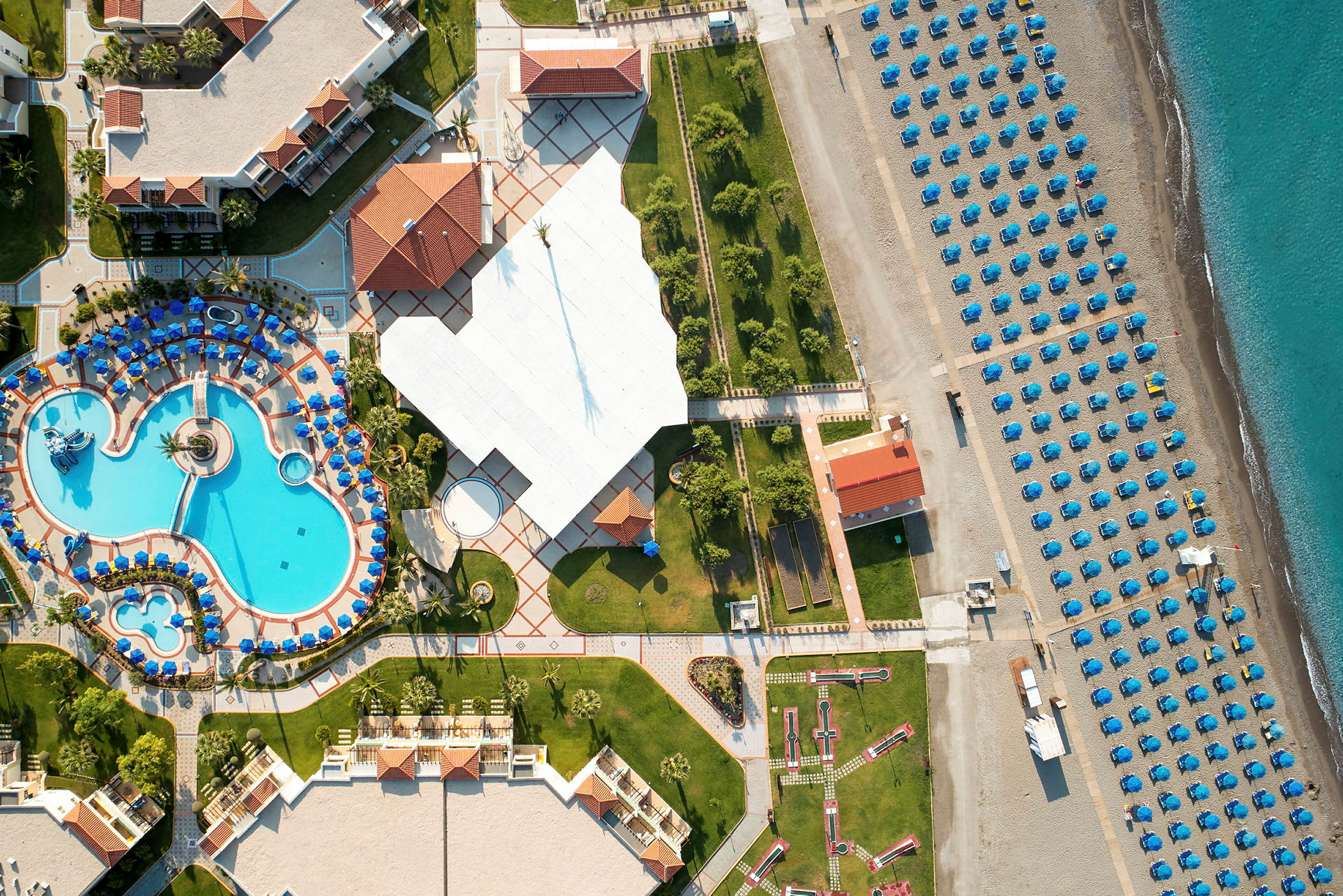Luftfoto der viser et feriested med poolområde, grønne områder og en strand med opstillede parasoller. (AI-genereret tekst)