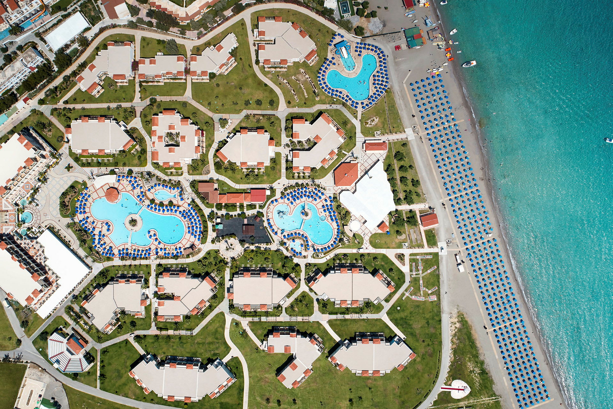 Fugleperspektiv af en resort med flere bygninger, poolområder og strand med parasoller. (AI-genereret tekst)