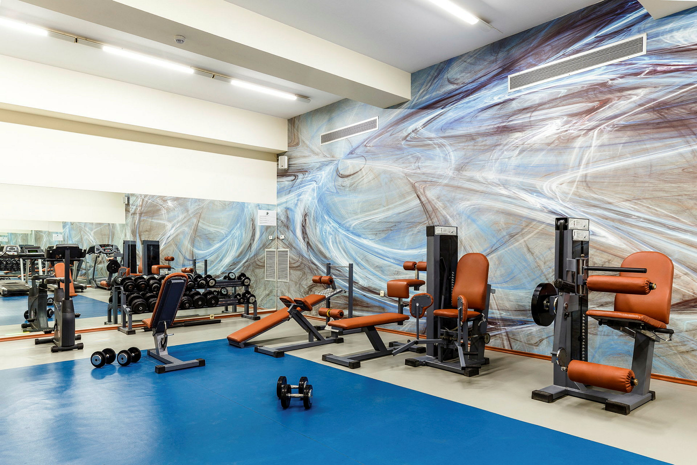 Billede af et fitnesscenter med træningsudstyr og en farverig vægmaleri. (AI-genereret tekst)