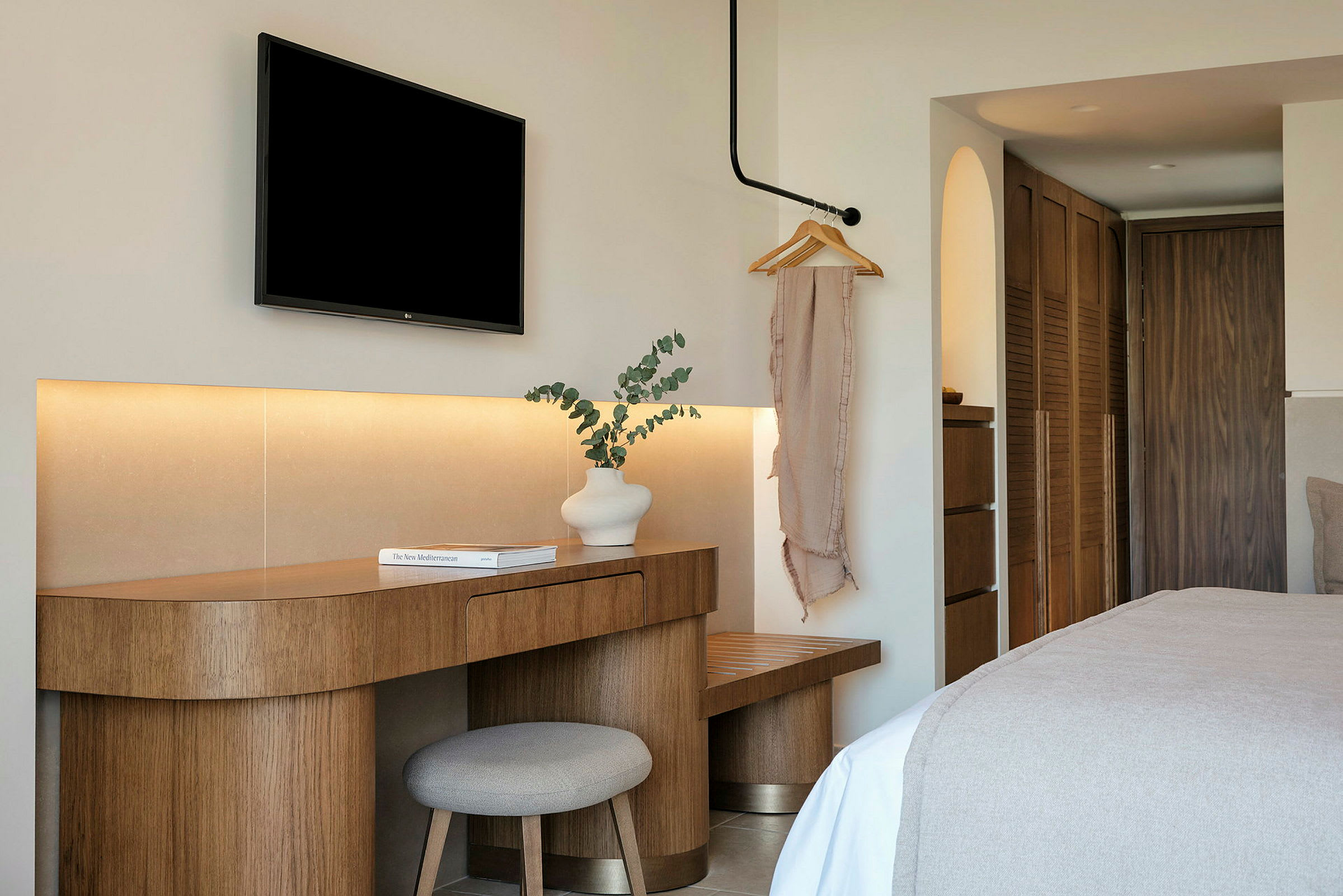 Et hotelværelse med moderne indretning. Et træbord med en hvid vase og en bog står ved siden af en stol. Et sort TV er monteret på væggen ovenover. (AI-genereret tekst)