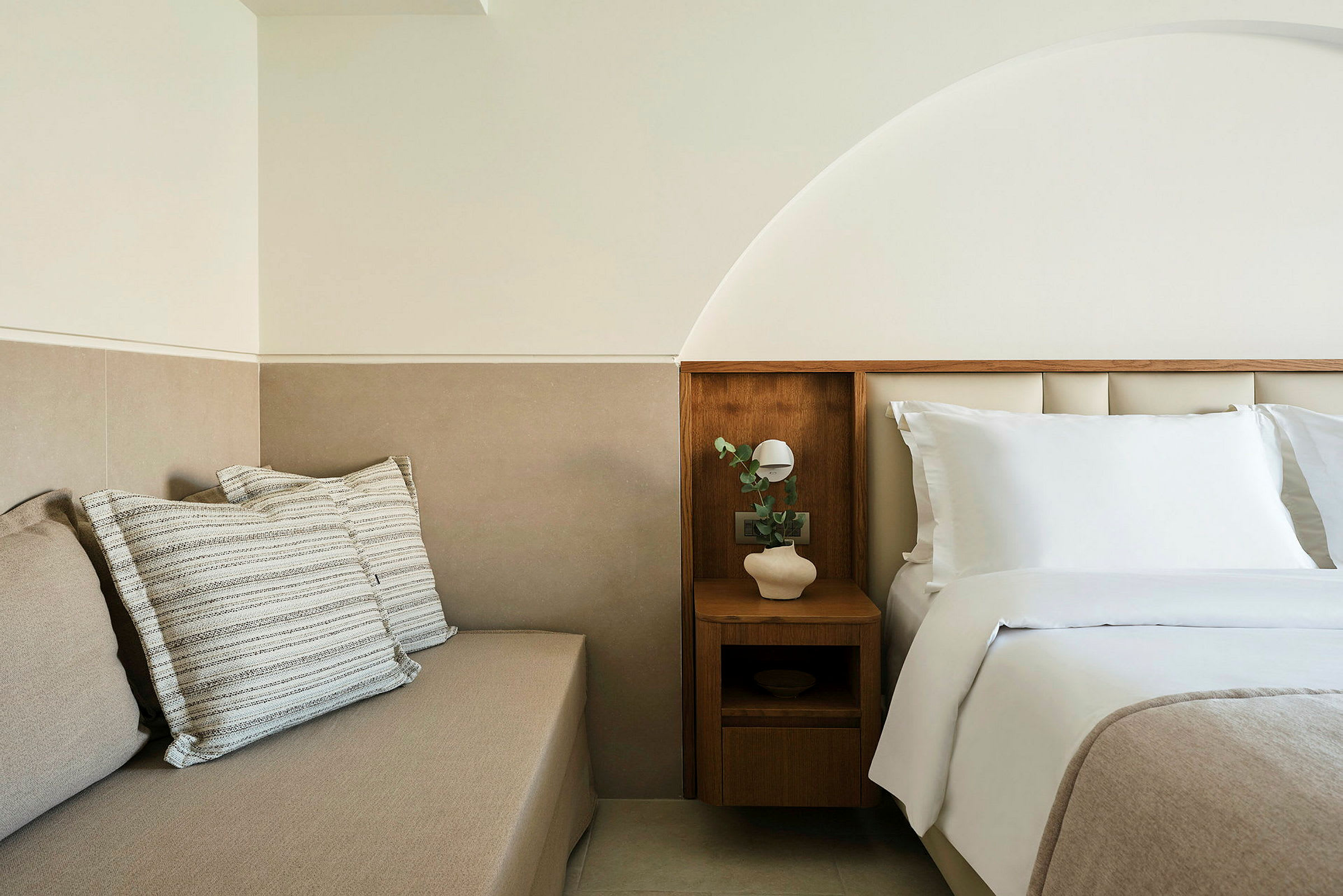 Et lyst og moderne hotelmiljø med en opredt seng og et siddemøbel med puder. (AI-genereret tekst)