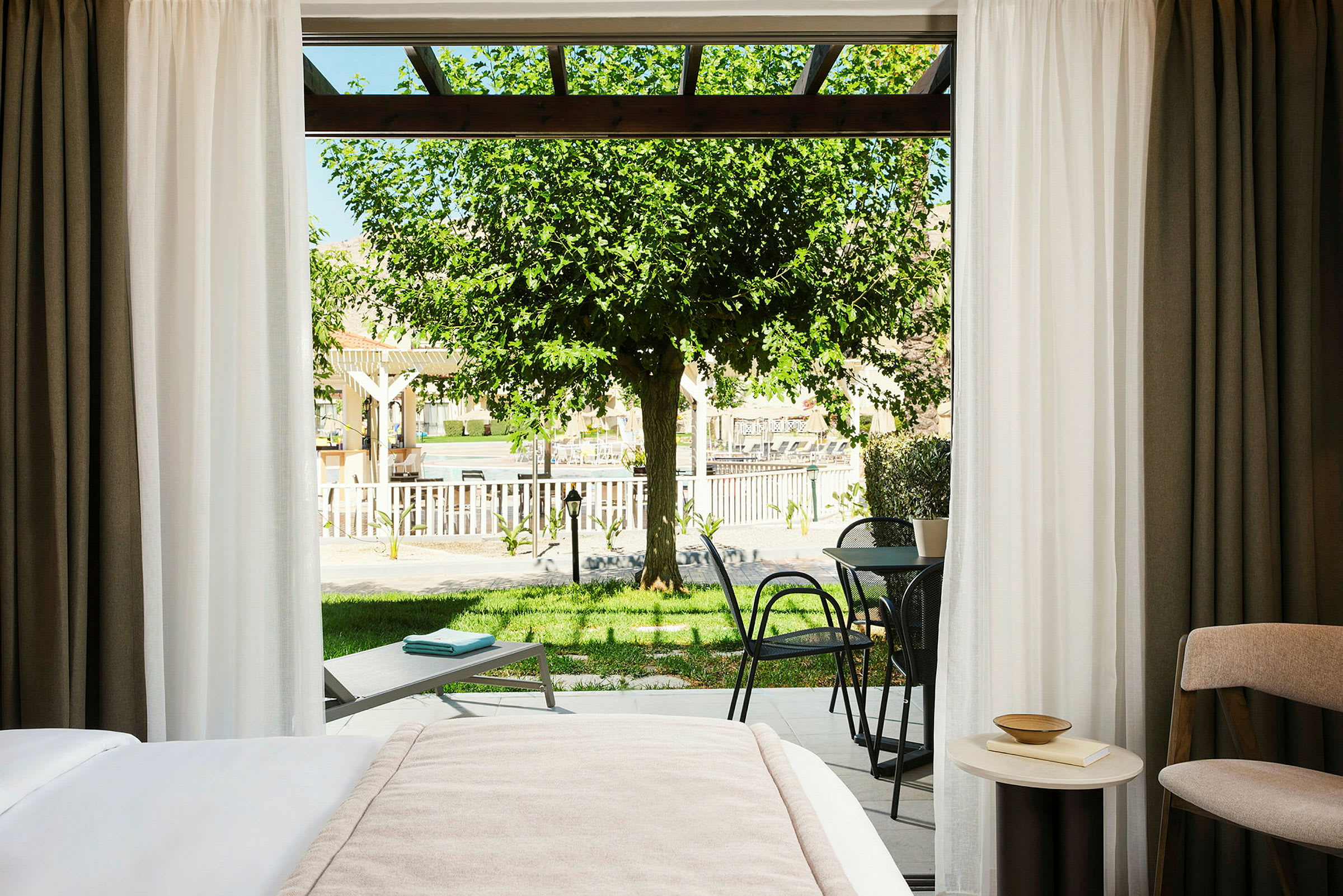 Udsigt fra et hotelværelse med en opredt seng, et bord med en bog og skål, en terrasse med stole og bord. Udenfor ses en græsplæne, et solsengsområde og et træ. (AI-genereret tekst)