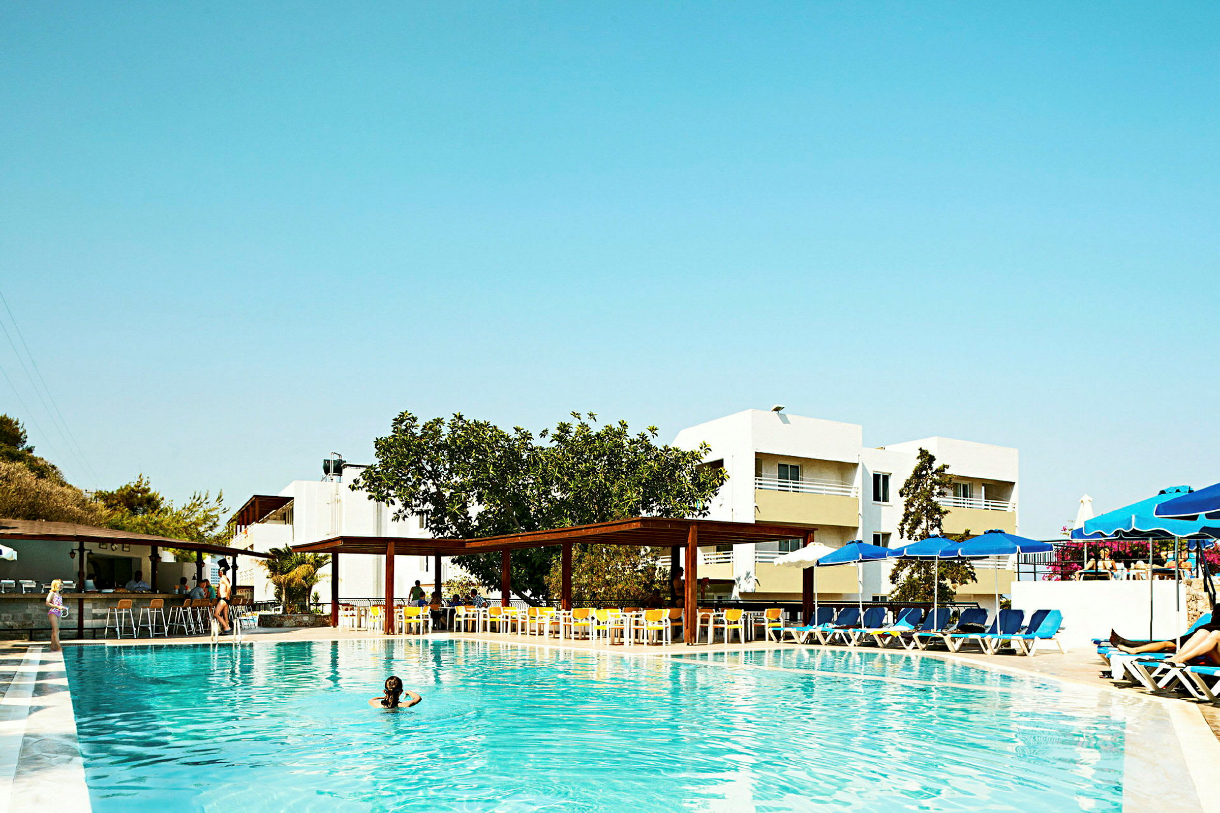 En udendørs swimmingpool med liggestole, parasoller og bygninger i baggrunden. (AI-genereret tekst)