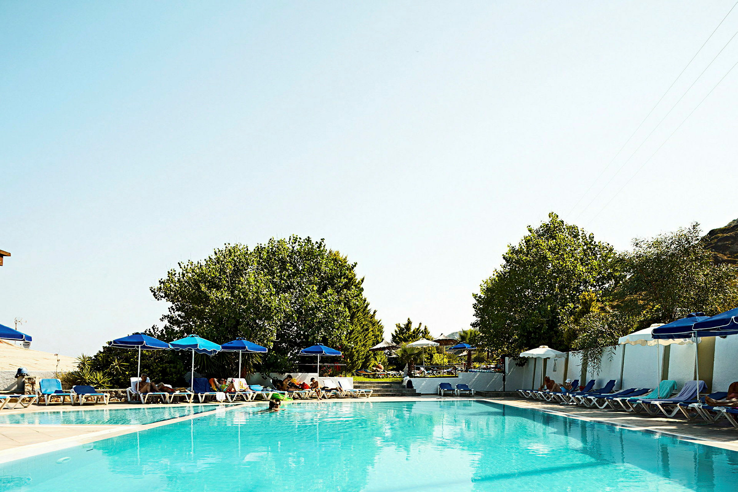 En udendørs swimmingpool omgivet af liggestole og parasoller med træer i baggrunden. (AI-genereret tekst)