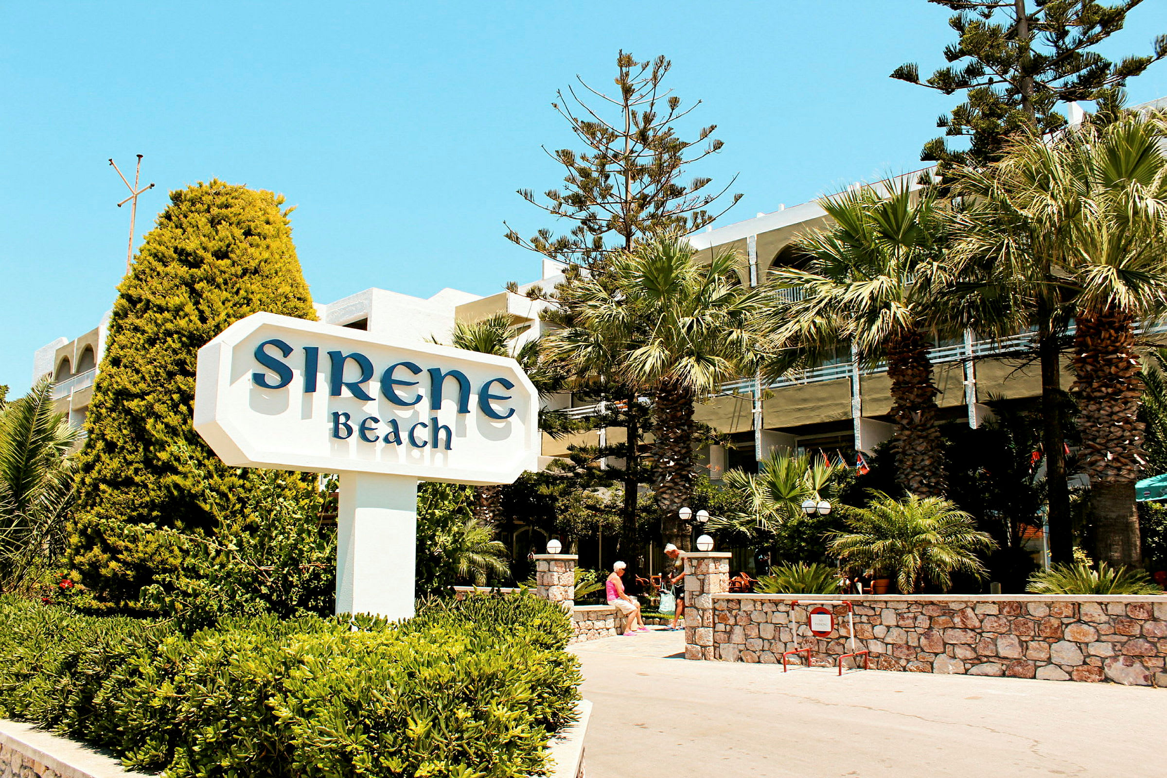Skiltet med teksten 'Sirene Beach' foran et hotel med palmer og beplantning. (AI-genereret tekst)