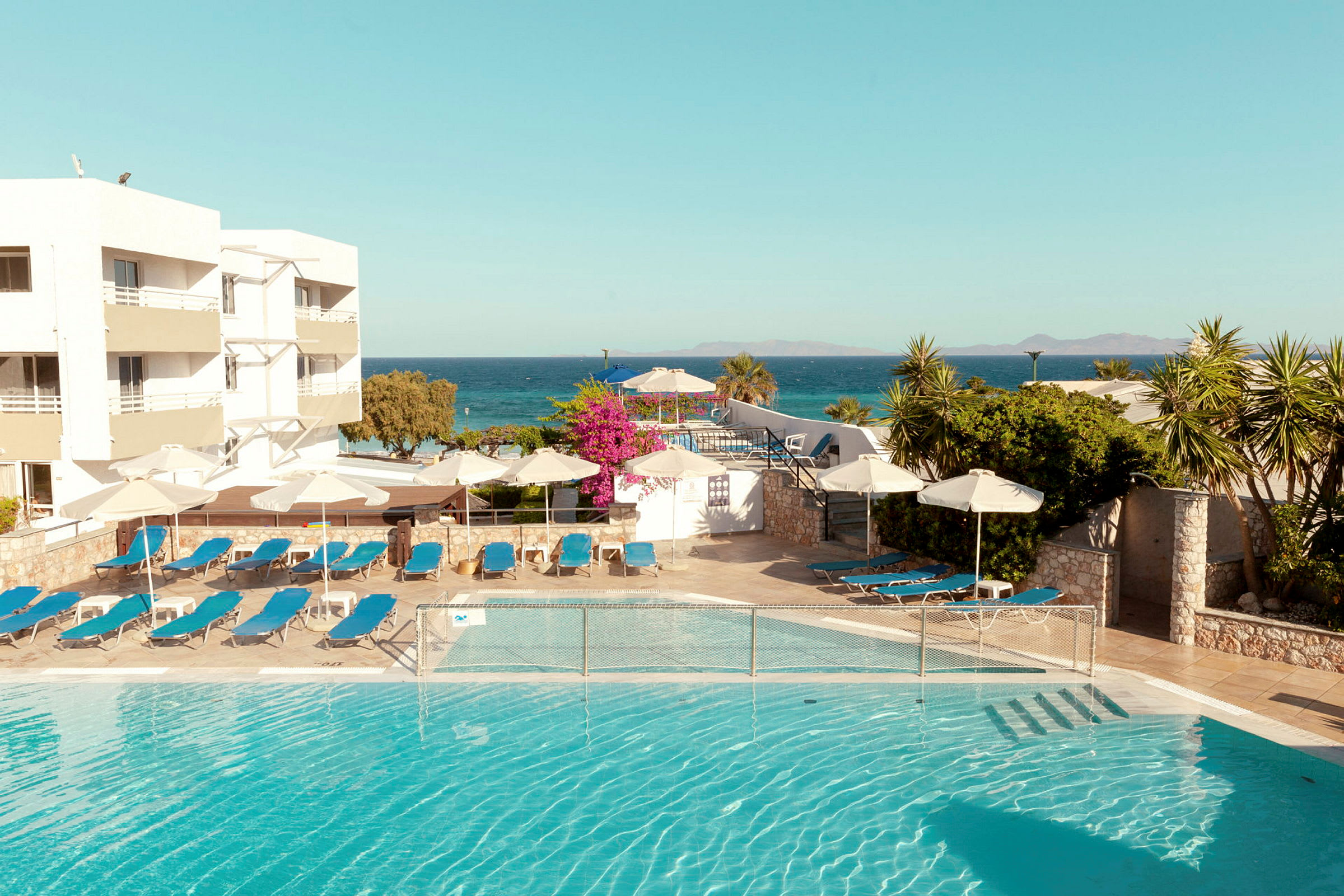 En udendørs pool med liggestole og parasoller ved siden af et hotel ved havet. (AI-genereret tekst)