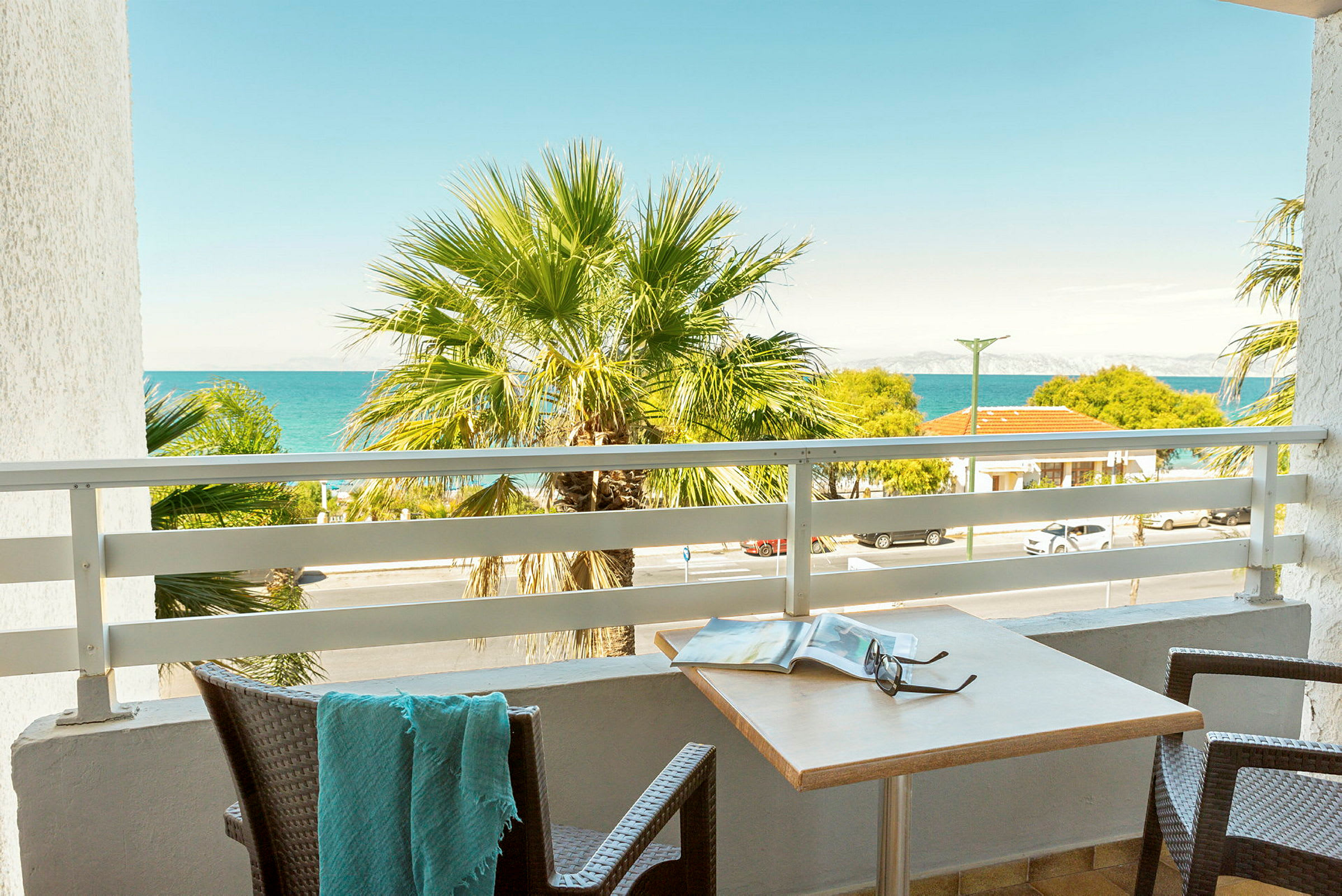 En balkon med udsigt over en palme, havet og nabobygninger. (AI-genereret tekst)