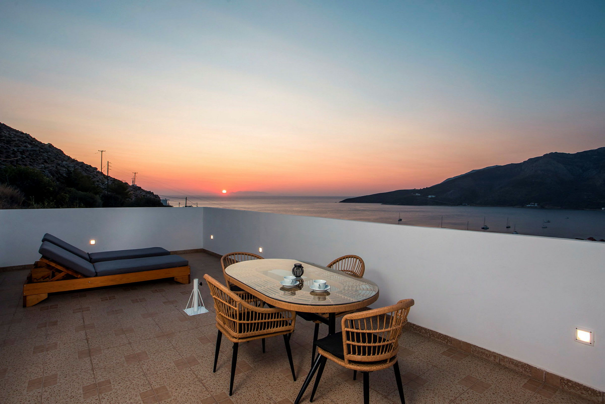 Udendørs terrasse med bord, stole og liggestol foran havudsigt ved solnedgang. (AI-genereret tekst)