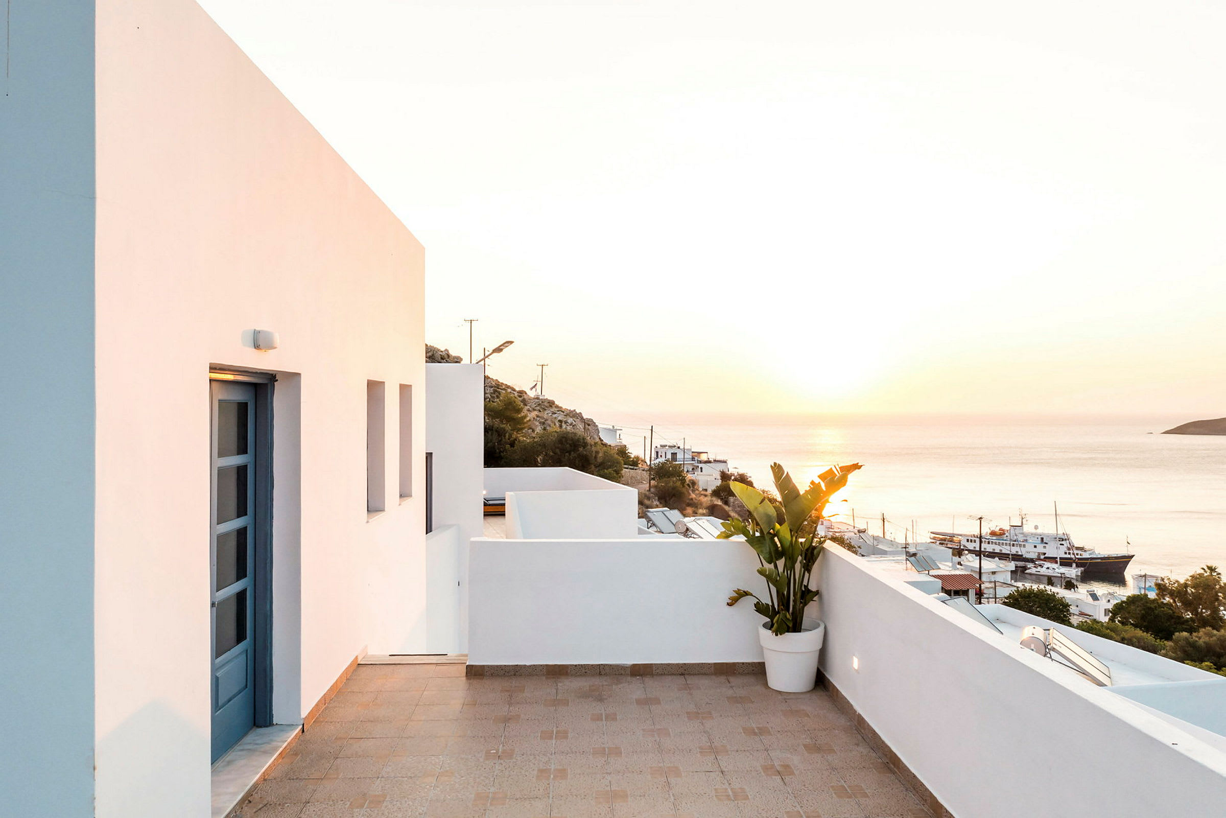 Terrasse med hvid husfacade og havudsigt ved solnedgang, en plante i en potte står ved rækværket. (AI-genereret tekst)