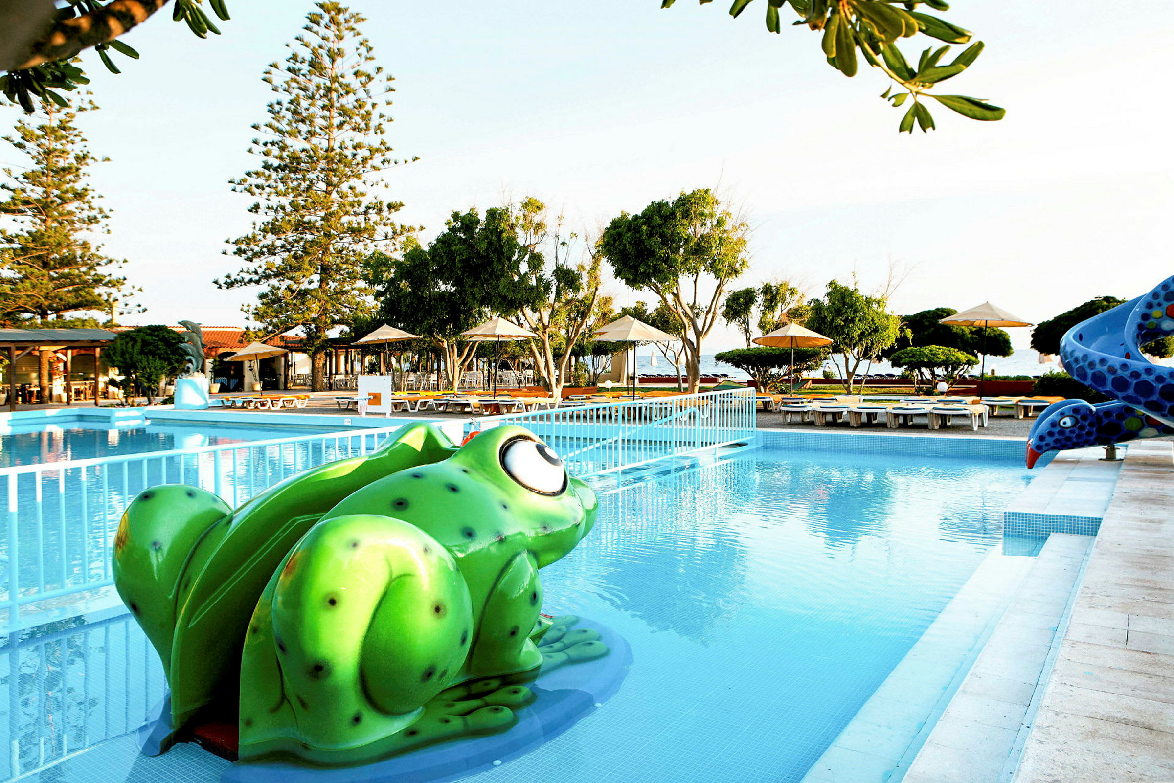 Udendørs swimmingpool med en grøn frøformet vandrutsjebane og omkringliggende liggestole under parasoller. (AI-genereret tekst)