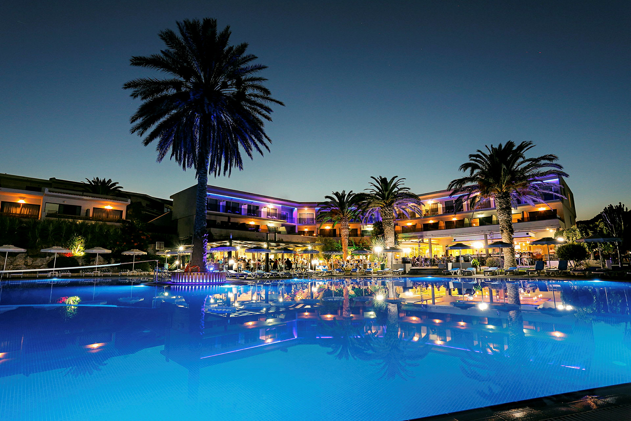 En oplyst udendørs pool foran et hotel omgivet af palmer under en mørk himmel. (AI-genereret tekst)
