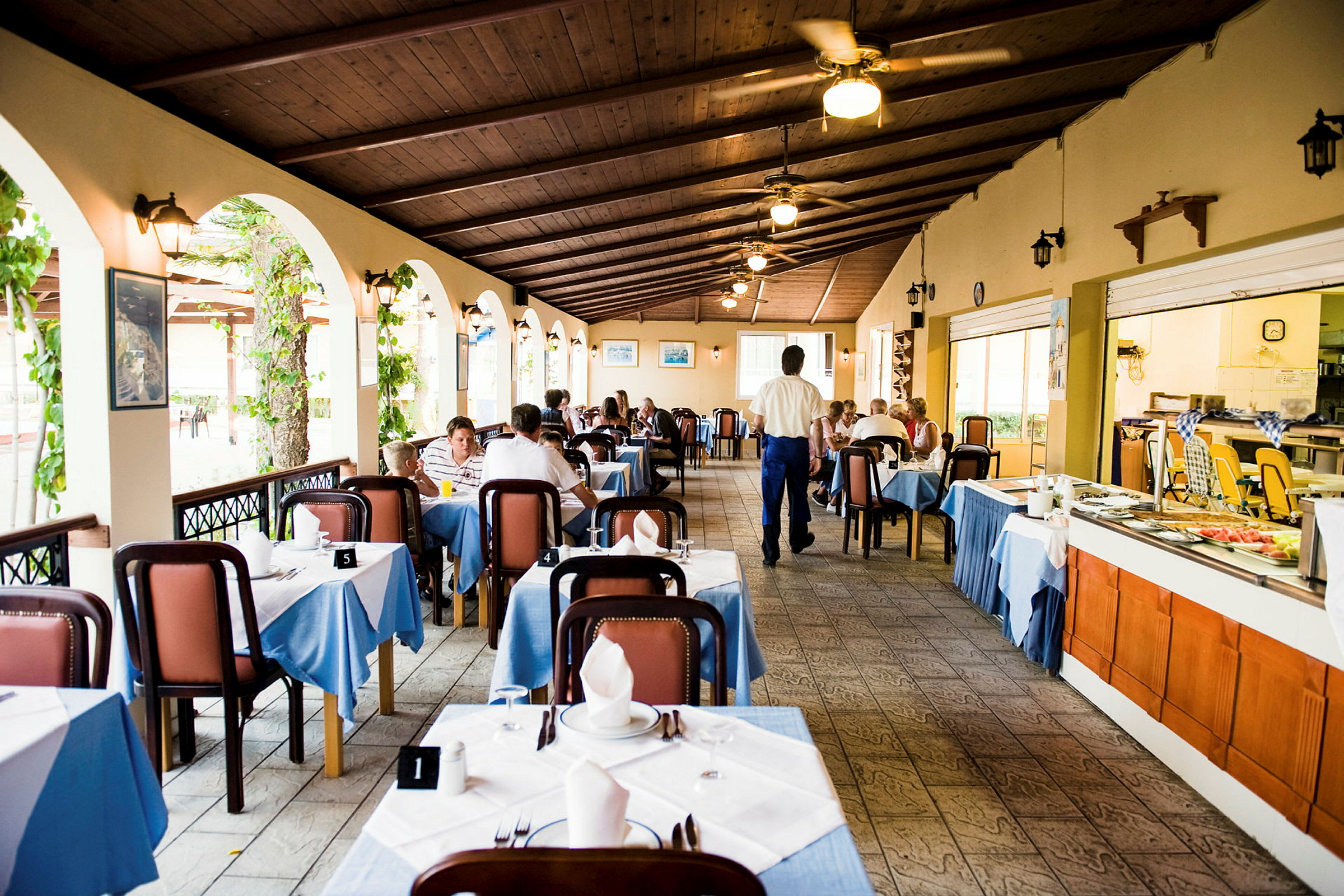 En restaurant med dækkede borde og gæster, der spiser middag i en hyggelig atmosfære. Vinduesåbninger med grønne planter på den ene side. (AI-genereret tekst)