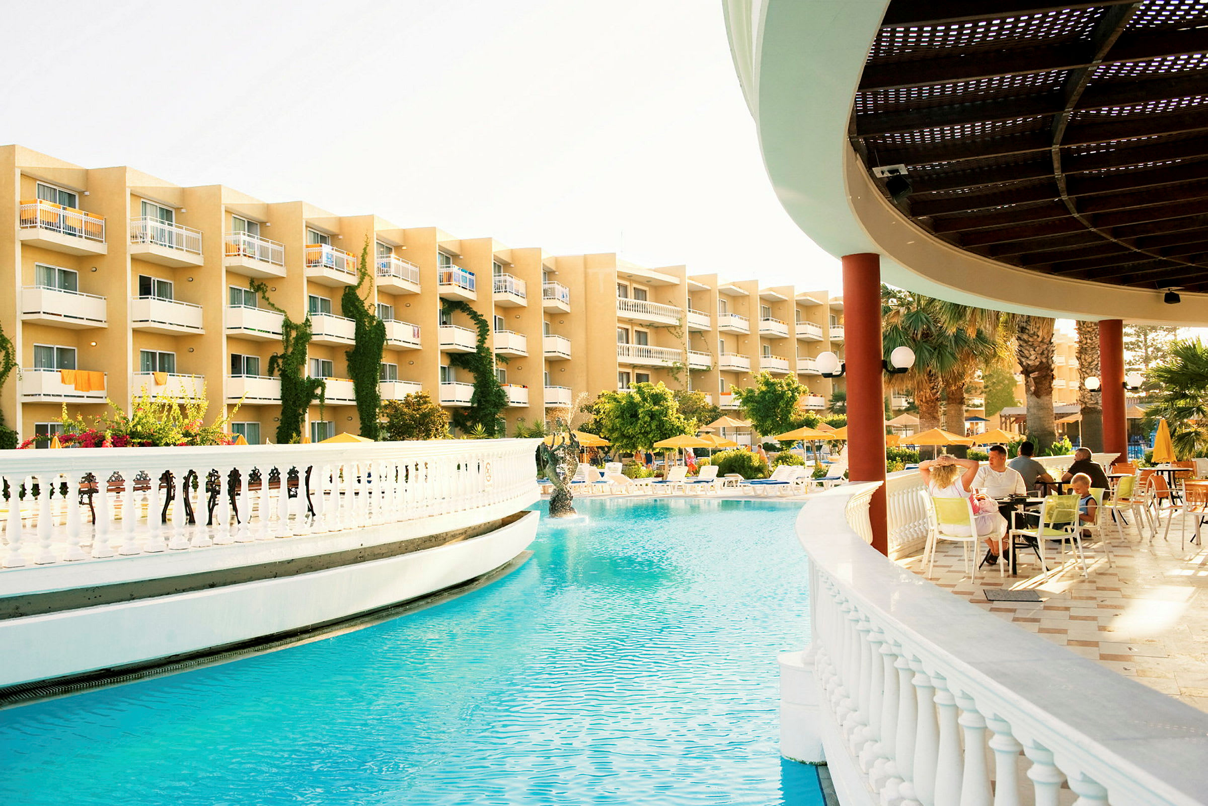 En hotelpool omgivet af solsenge og parasoller, med en bygning i baggrunden og et overdækket område med siddepladser. (AI-genereret tekst)