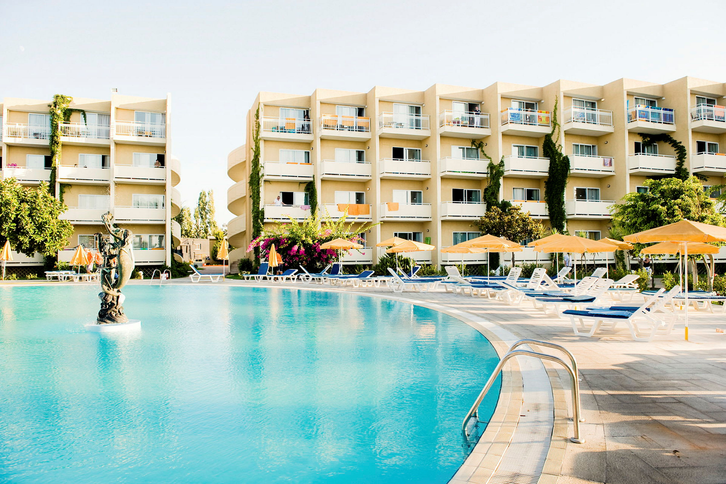 Et udendørsbillede, der viser en stor pool med en statue i midten, omgivet af liggestole og parasoller foran en hotelbygning. (AI-genereret tekst)