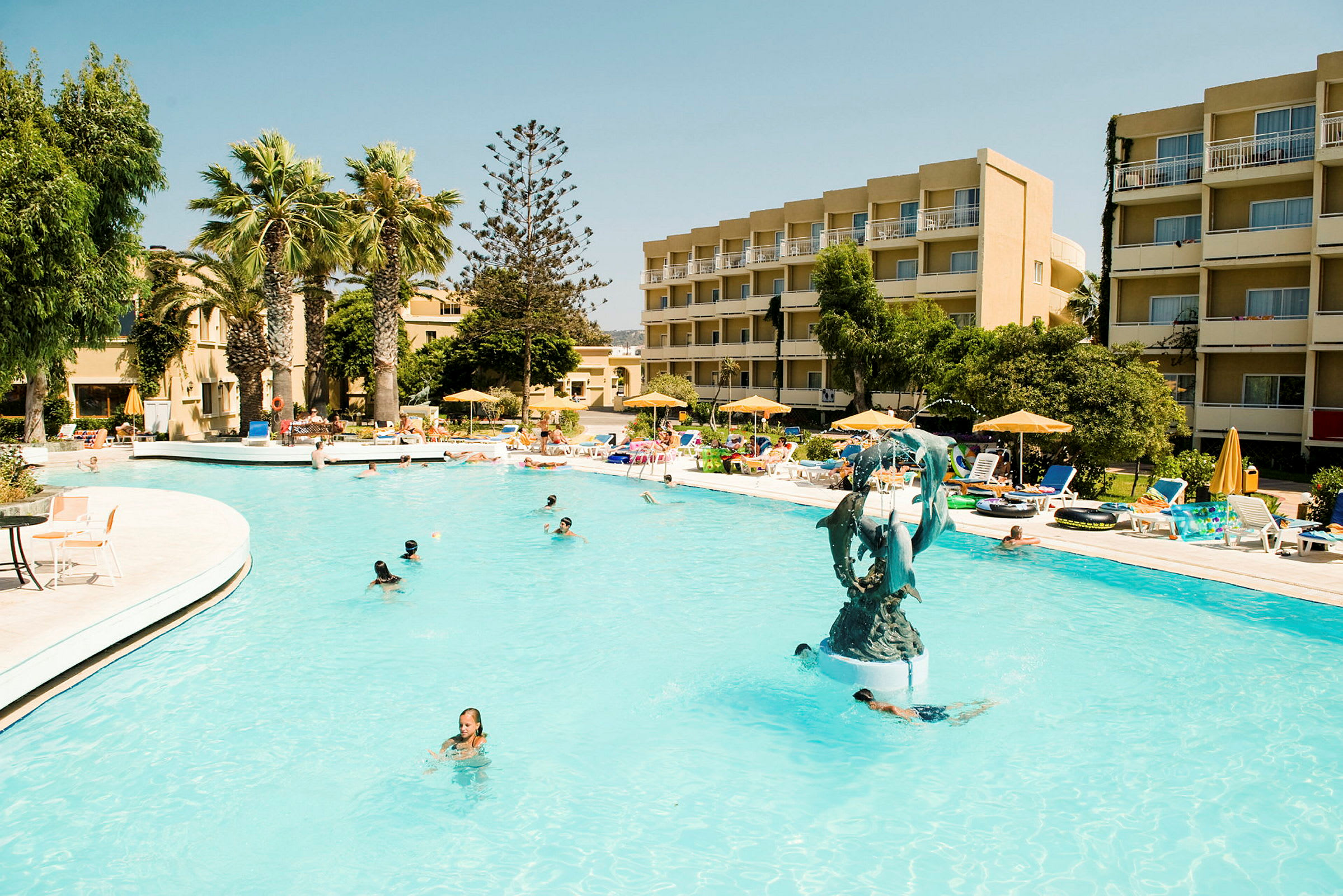 En udendørs swimmingpool omgivet af liggestole, parasoller og en bygning. I poolen er der personer, der svømmer, samt en fontæne med en delfinskulptur. (AI-genereret tekst)