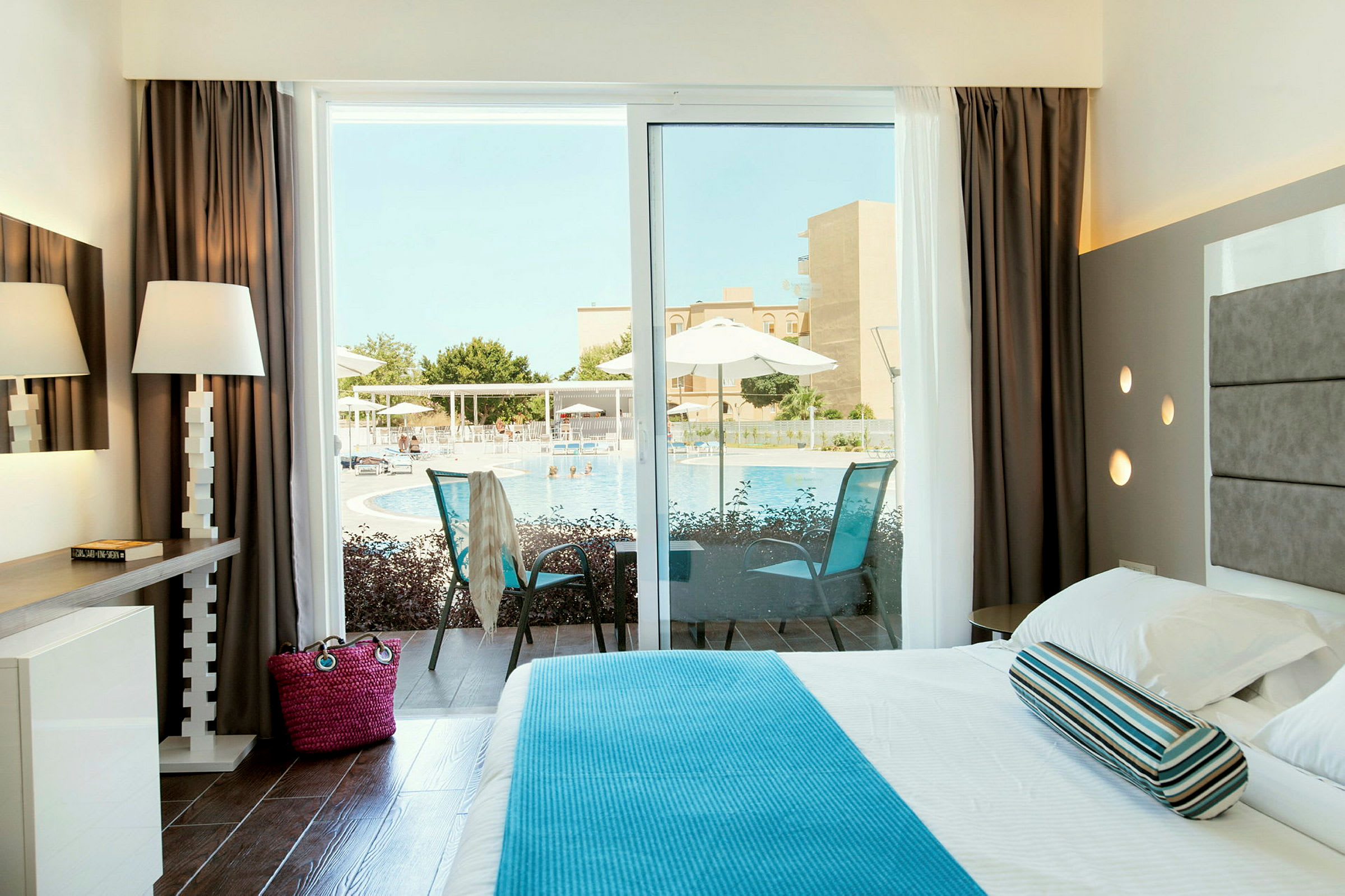 Et lyst hotelværelse med udsigt over en pool gennem en glasdør. (AI-genereret tekst)