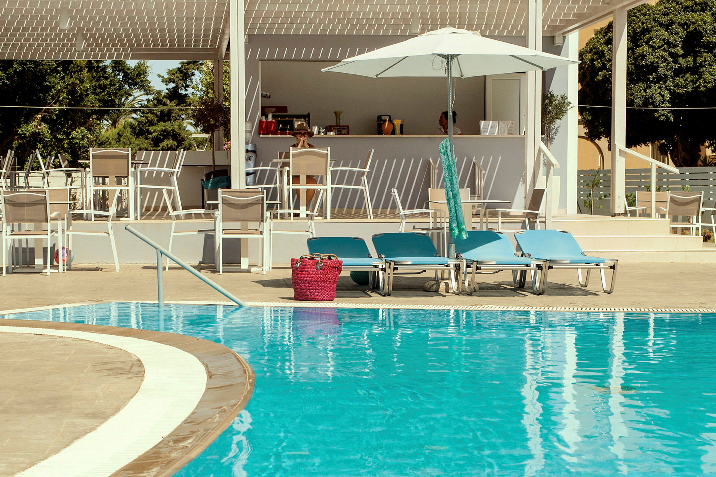 Billede af et poolområde med liggestole, parasol og en bar i baggrunden. (AI-genereret tekst)
