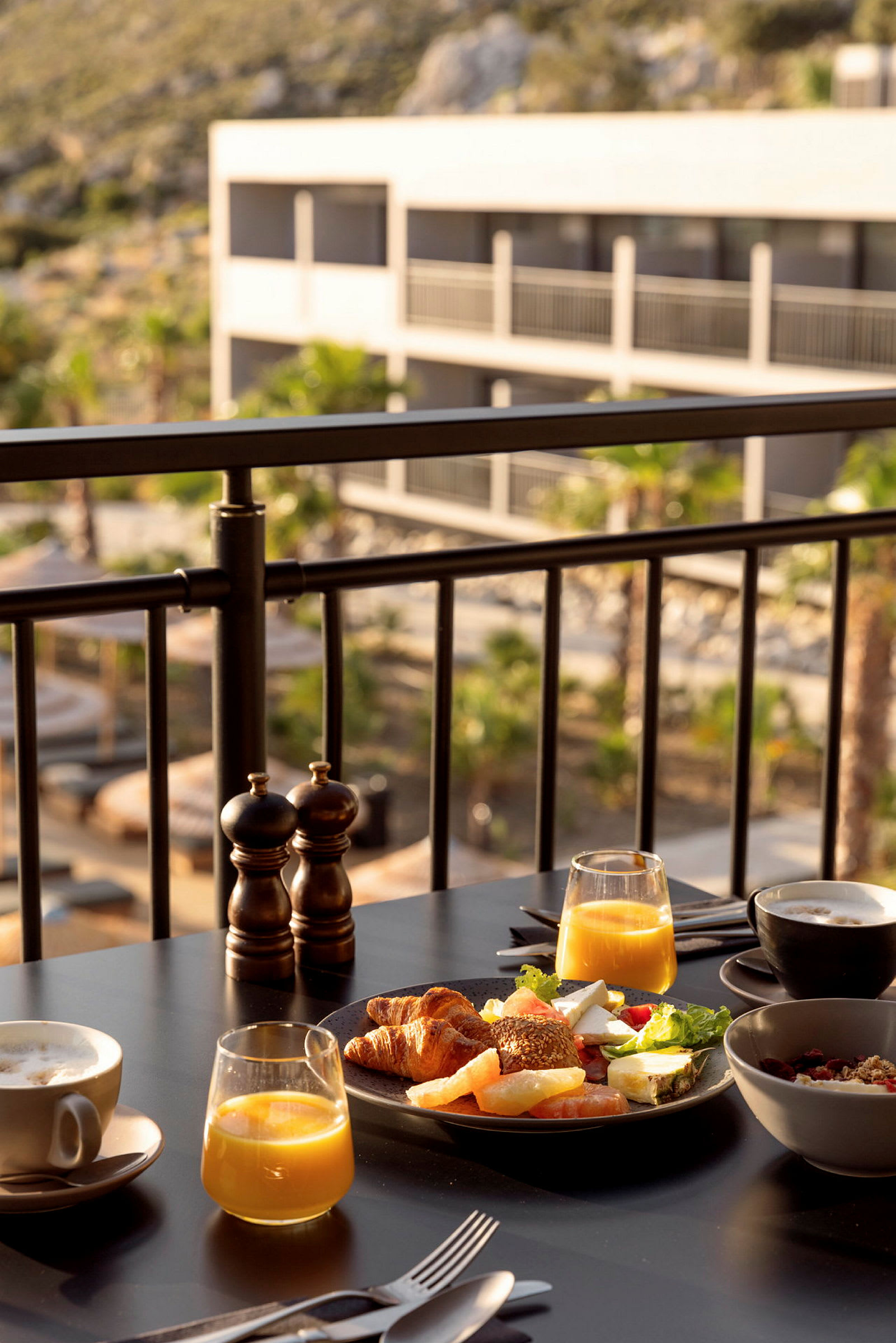Morgenmad med juice, kaffe og forskellige brød serveret ved et bord på en solterrasse med udsigt over en hotelbygning. (AI-genereret tekst)
