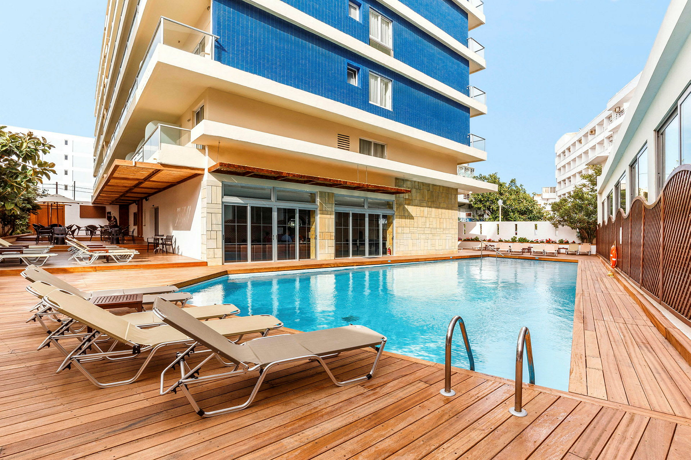 Udendørs pool med liggestole og hotelbygningen i baggrunden. (AI-genereret tekst)