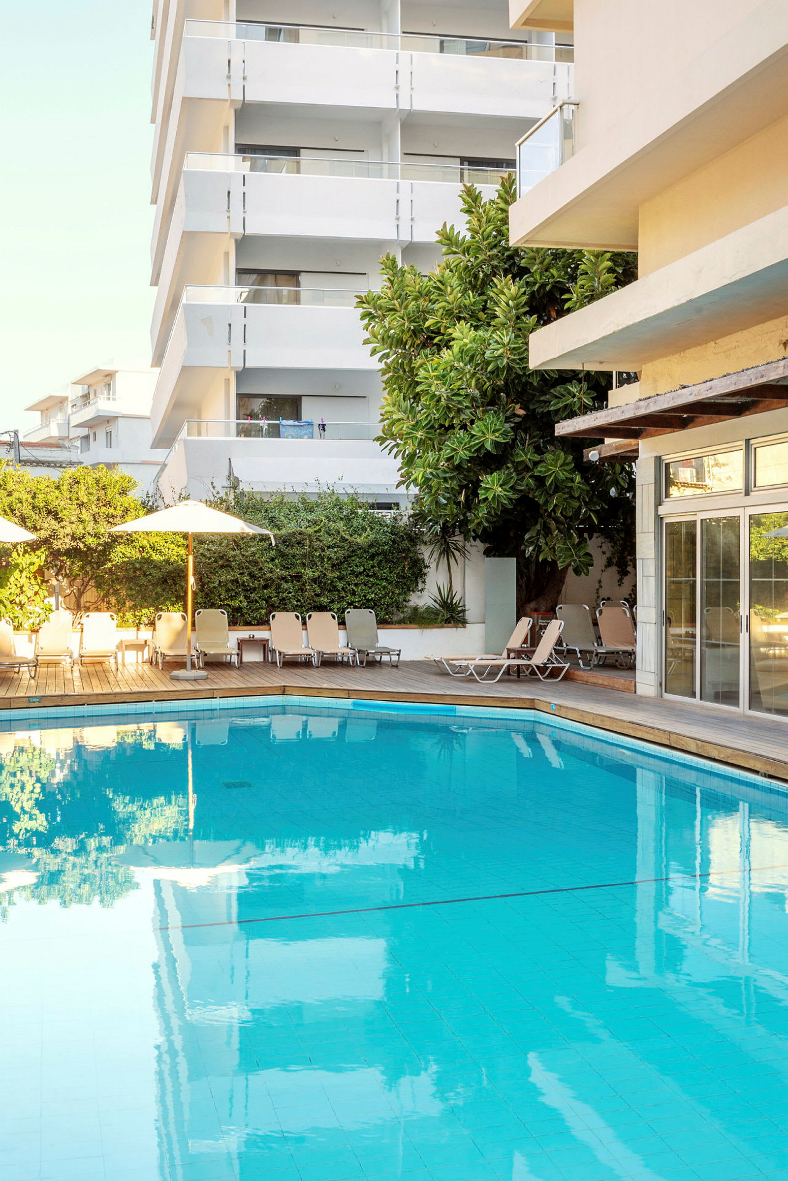 Udendørs pool med liggestole og hotelbygning i baggrunden. (AI-genereret tekst)