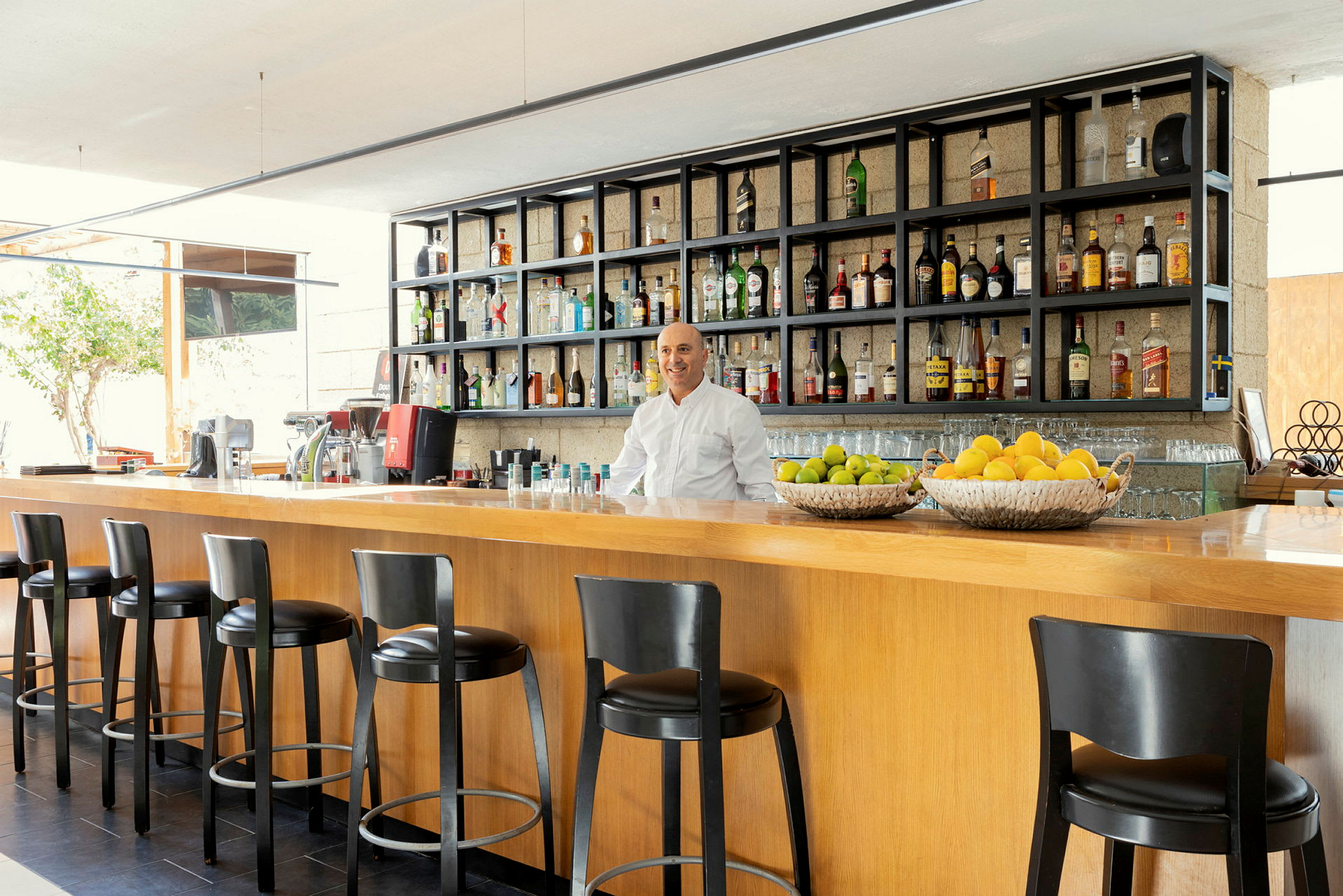 En moderne bar med høje stole ved en bardisk i træ, hylder fyldt med flasker og frugtkurve med citrusfrugter. (AI-genereret tekst)