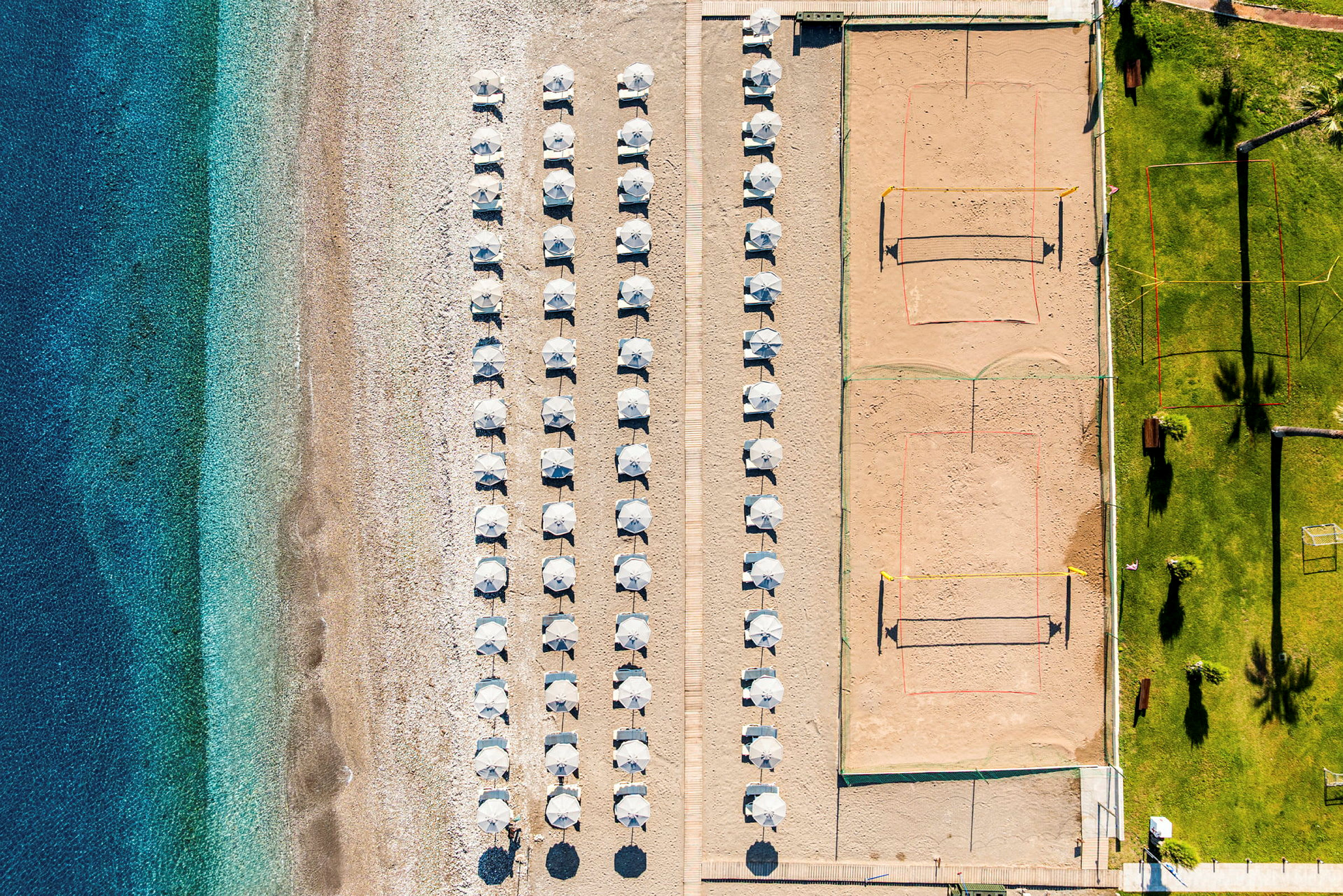 Luftfoto af en strand med parasoller opstillet i rækker, en volleyballbane på sand ved siden, og en græsplæne med palmer og siddepladser. (AI-genereret tekst)