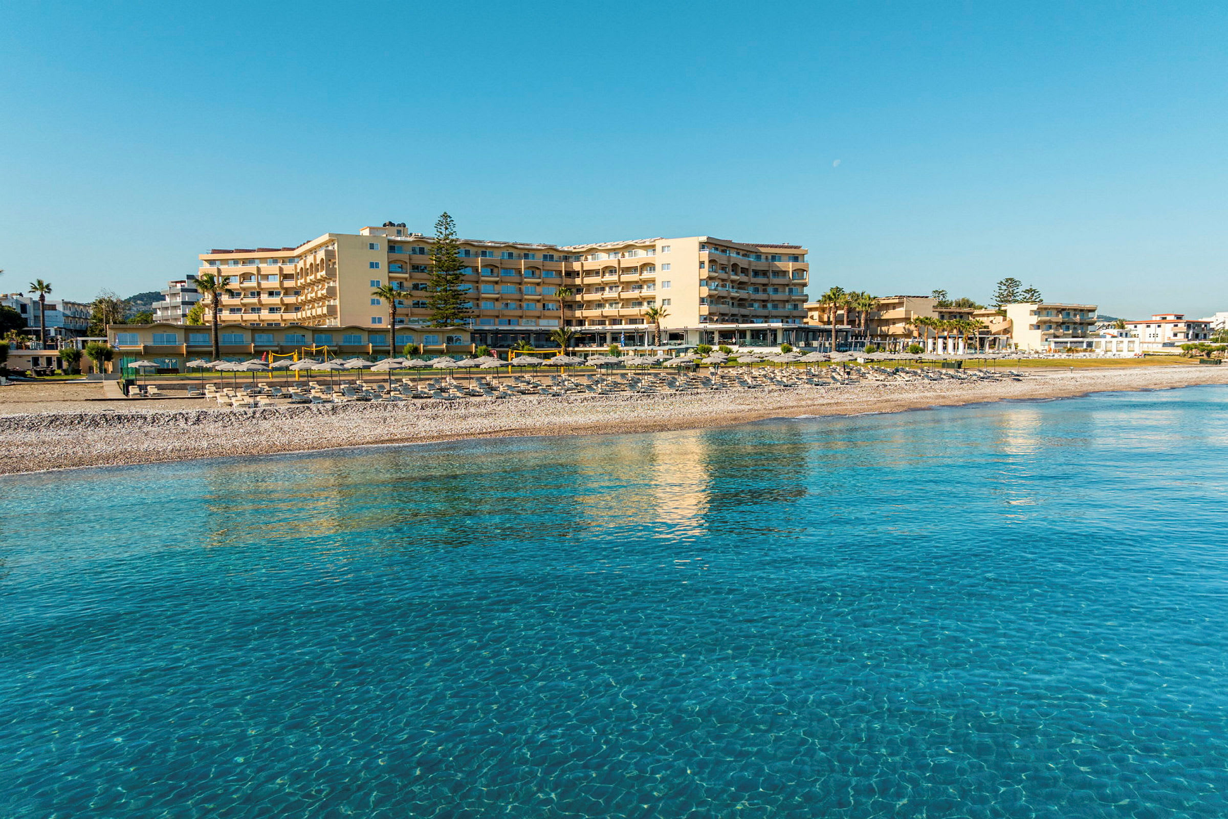 Et hotel ved stranden med klart blåt vand i forgrunden. (AI-genereret tekst)