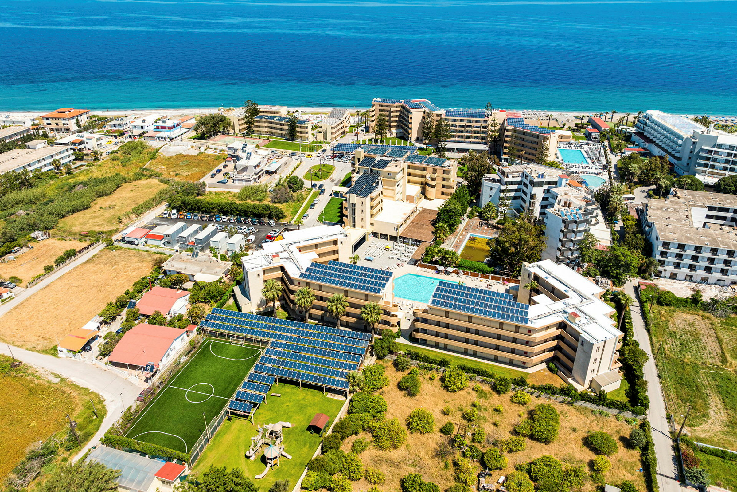 Luftfoto af et resortområde med hoteller, swimmingpools, grønne arealer og en legeplads, beliggende tæt på stranden og havet. (AI-genereret tekst)