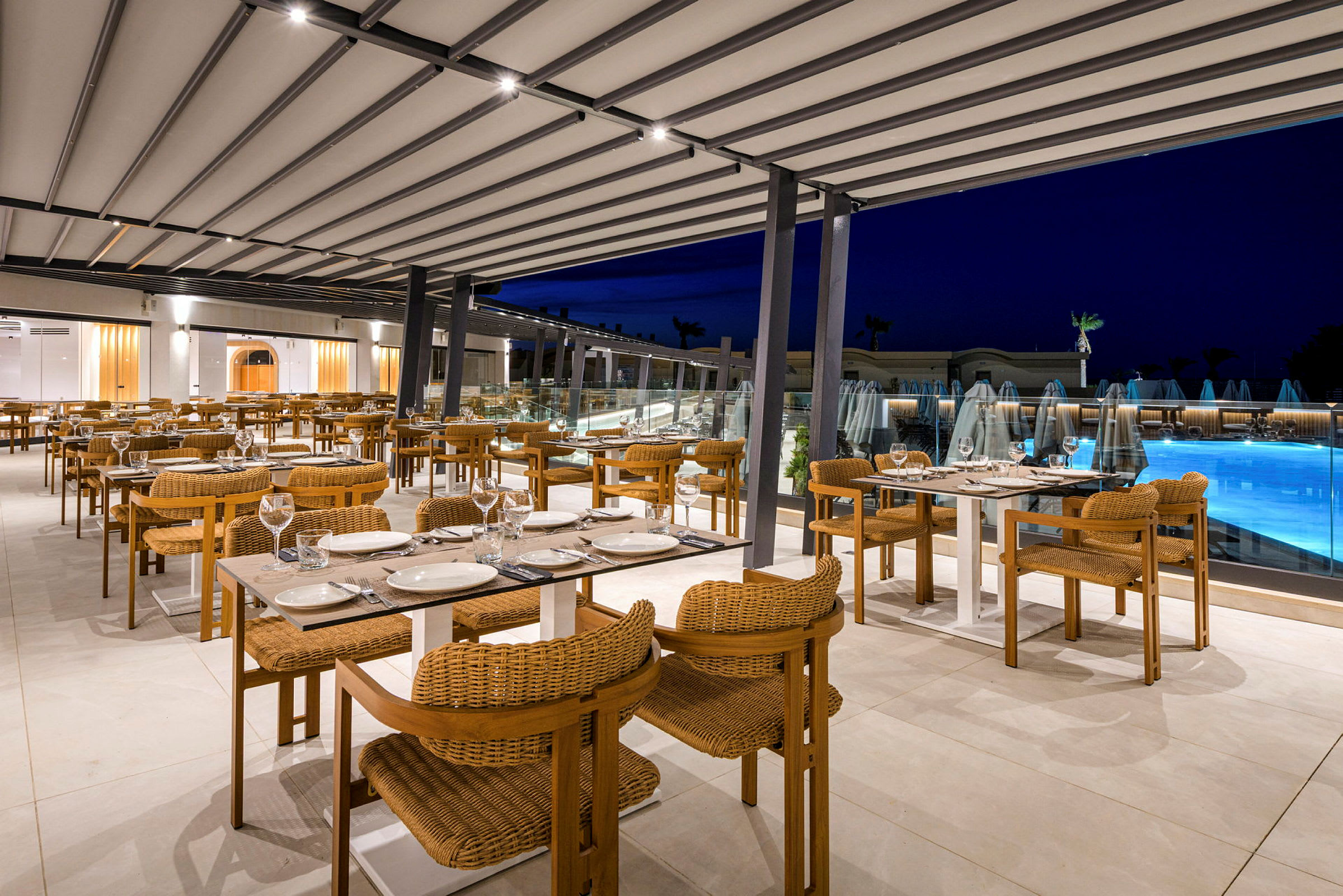 En restaurant med udendørs siddepladser, dækkede borde og udsigt til en oplyst pool. (AI-genereret tekst)