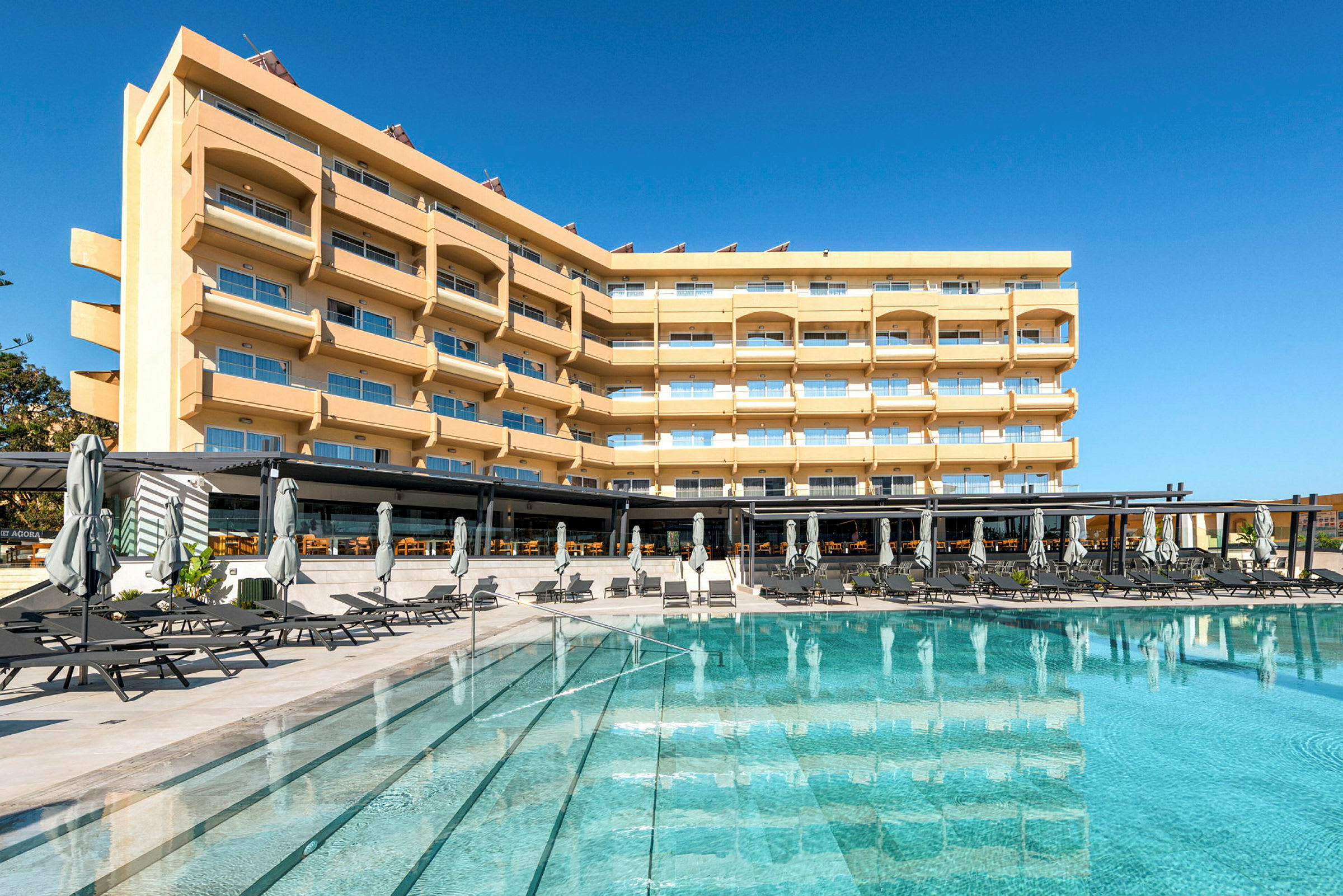 En hotelbygning med flere etager, balkoner og store vinduer, foran en swimmingpool med liggestole og parasoller. (AI-genereret tekst)