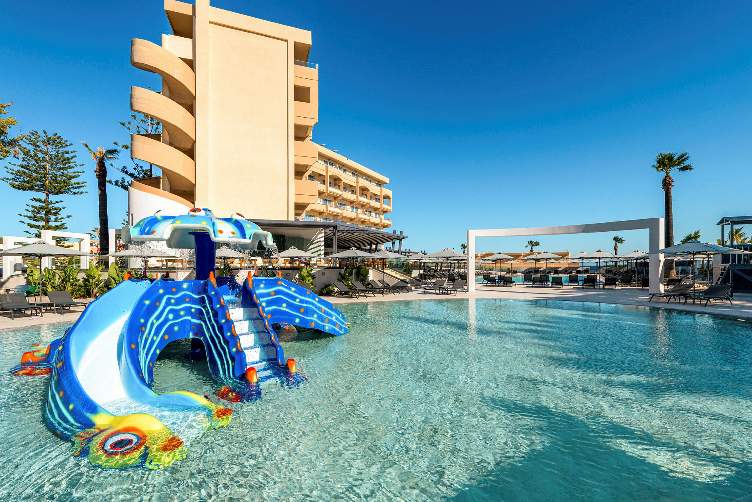 Billede af en pool med en sjov rutsjebane formet som en blæksprutte, omgivet af liggestole og et hotel i baggrunden. (AI-genereret tekst)