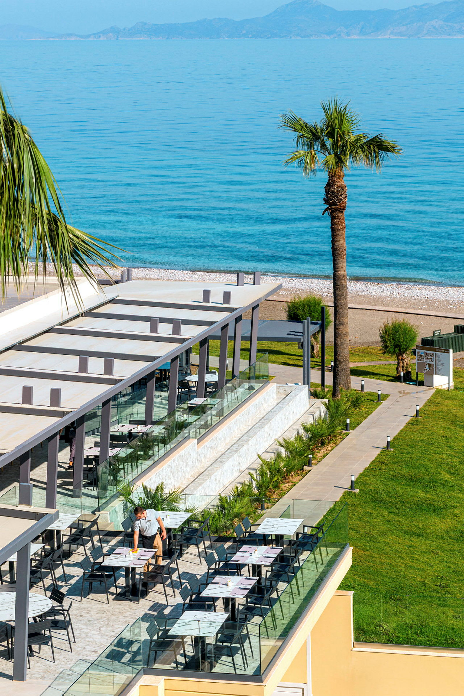 Udendørs terrasse med borde og stole, tilknyttet en bygning med udsigt til en strand med havet i baggrunden. (AI-genereret tekst)