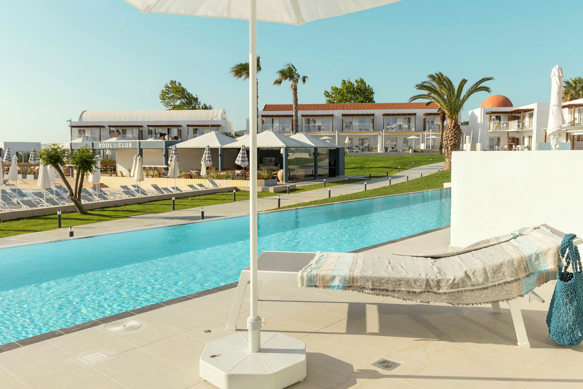 En solseng med et tæppe ved kanten af en pool, med en parasol i forgrunden og hotelbygninger i baggrunden. (AI-genereret tekst)