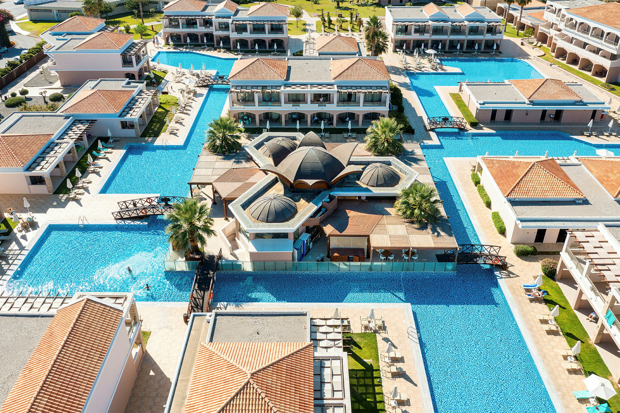 Luftfoto af hotelområdet med omkringliggende pools, bygninger og grønne områder. (AI-genereret tekst)